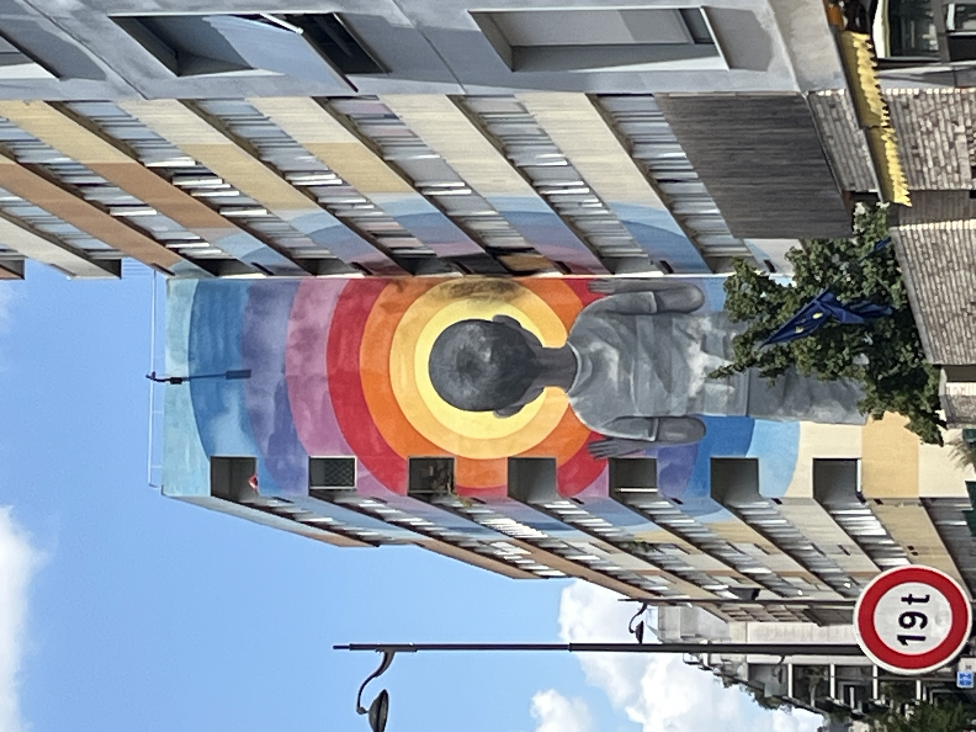 A mural of a young boy with his back to the viewer is painted on the side of a tall, multi-story building. The boy is rendered in grayscale and appears to be looking up at a series of concentric circles in vibrant rainbow colors. The building itself is painted with broad strokes of blue, pink, and yellow, with visible windows and balconies. To the left of the building, a tall lamppost stands on a sidewalk. A circular traffic sign with "19t" inside it is visible in the foreground, also to the left. Adjacent to the mural, another building with a more traditional facade of beige and brown vertical stripes, punctuated by windows, is partially visible. In front of the buildings, a cluster of green trees and a blue flag with yellow stars are present. The sky above is a clear blue with scattered white clouds. The scene appears to be a street view in an urban environment.