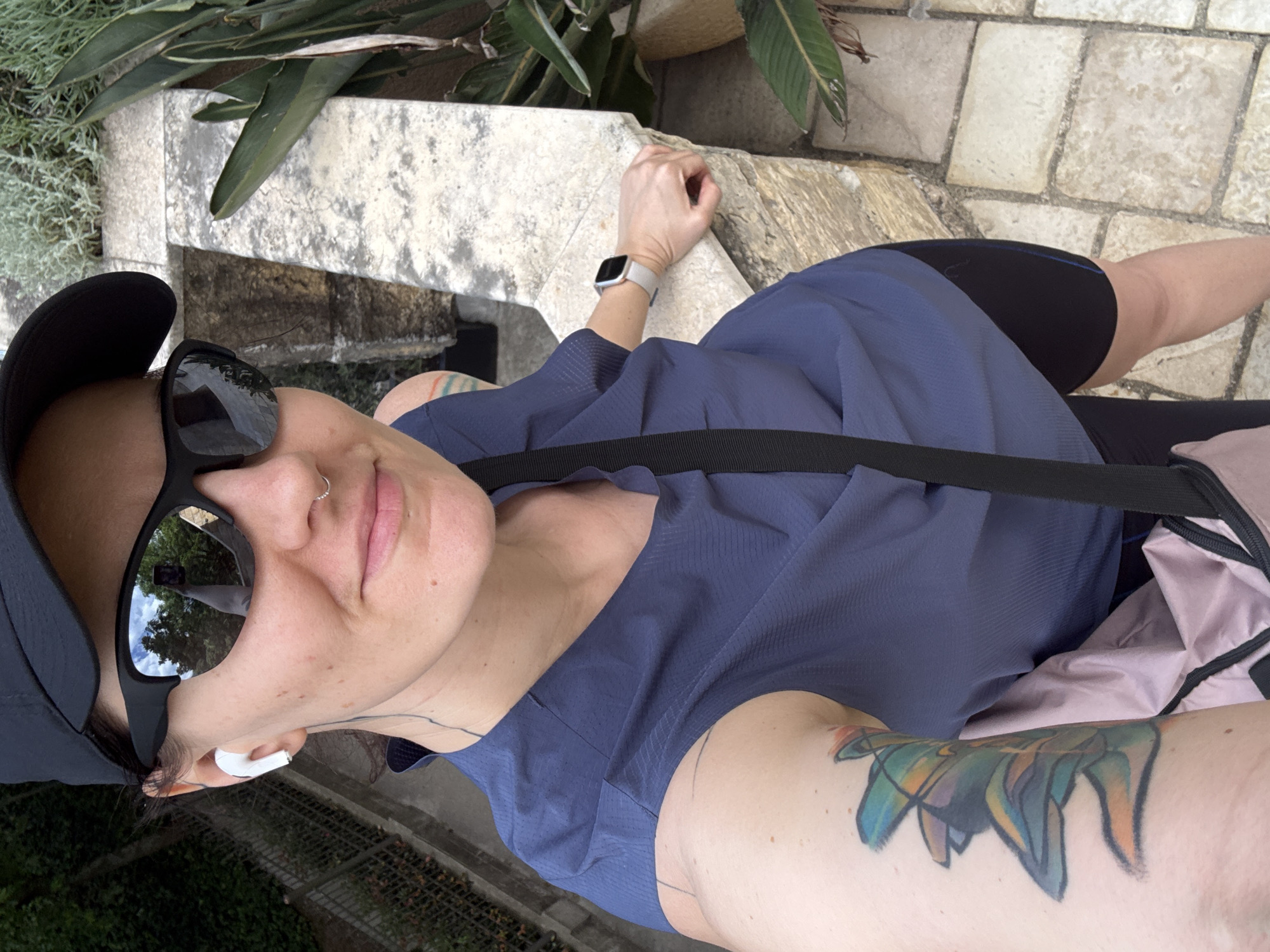 A selfie photograph taken outdoors depicts a person with light skin, wearing a black baseball cap, black sunglasses, and a blue short-sleeved shirt. The person has a nose ring and is wearing an earbud in their left ear. A black strap crosses their torso, suggesting a bag is worn crossbody. Visible tattoos are present on their arms. A silver smartwatch is on their left wrist. The surroundings include stone structures, greenery, and a stone-paved surface. The person is located at the Sveti Djordje area in the Boreti, Montenegro.