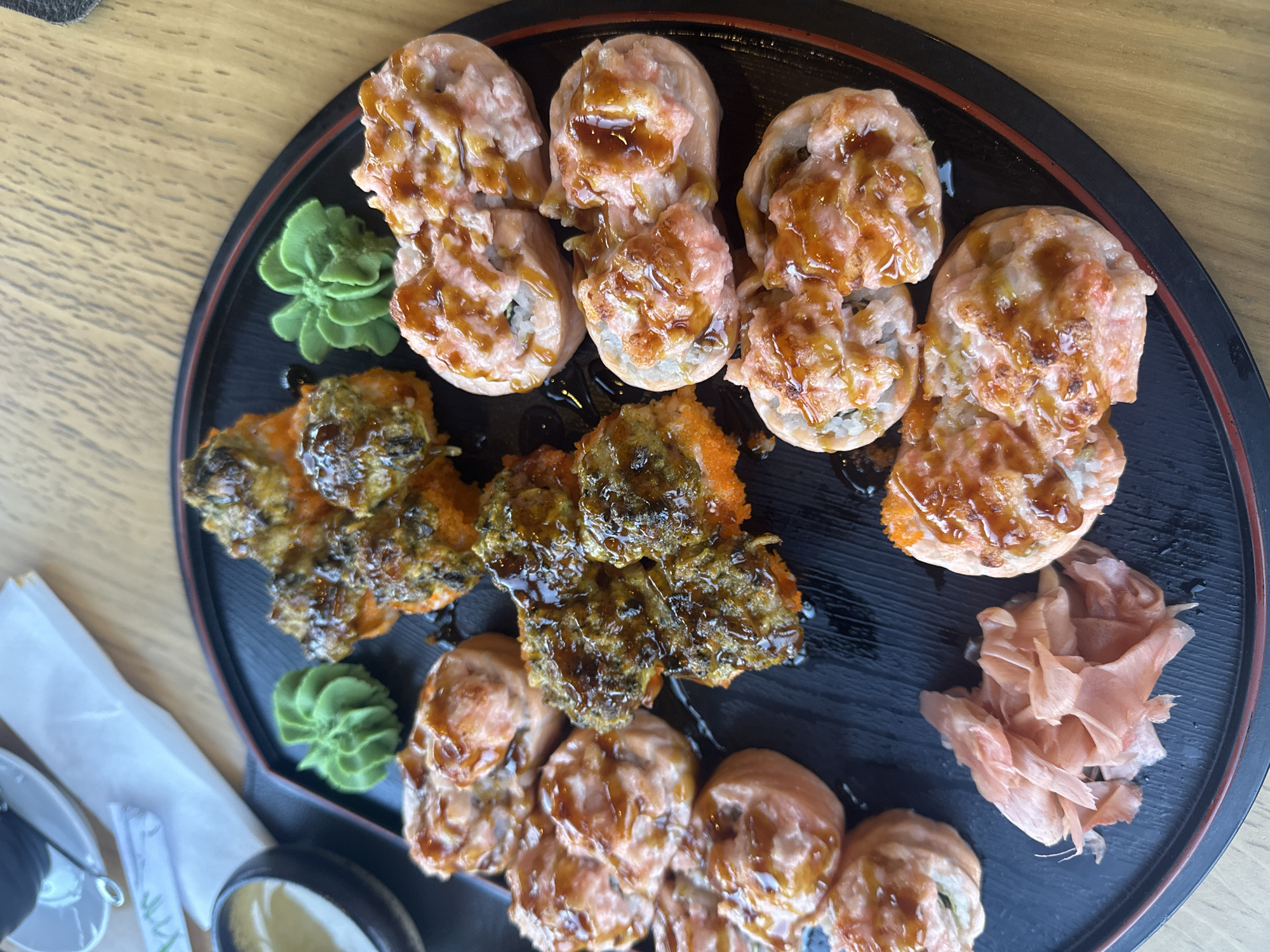 A black, round plate with a red rim sits on a wooden surface, holding an assortment of sushi. The sushi is arranged in three groupings: one consisting of pieces topped with a pink, sauce-drizzled mixture; another with pieces covered in a darker, textured topping; and a final one that resembles the first. Beside the sushi are small mounds of wasabi and pickled ginger. A container of dipping sauce and a pair of sunglasses can be partially seen on the left side of the image. The scene appears to be set in a restaurant in Riga, Latvia.