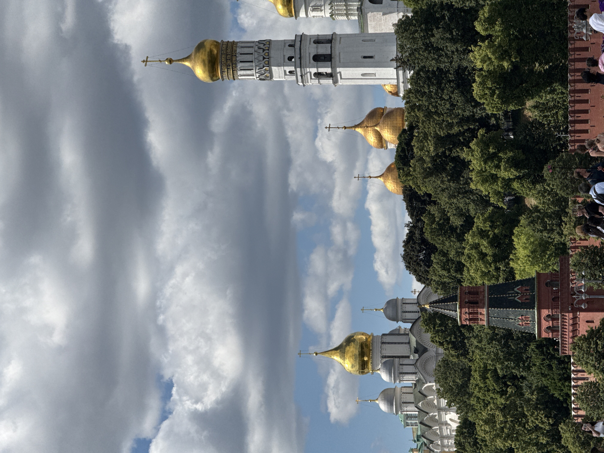 This image shows a wide shot of the Moscow Kremlin complex, dominated by its distinctive white bell towers and golden-domed churches. The Kremlin's architecture, characterized by its intricate stonework and ornate religious symbols, is set against a backdrop of lush green trees. A cloudy sky with patches of blue adds a dramatic contrast to the scene. In the foreground, the red brick walls of the Kremlin and a group of people are visible, hinting at the public accessibility of the area. The image captures the grandeur and historical significance of the Moscow Kremlin, a UNESCO World Heritage site and the official residence of the President of the Russian Federation.