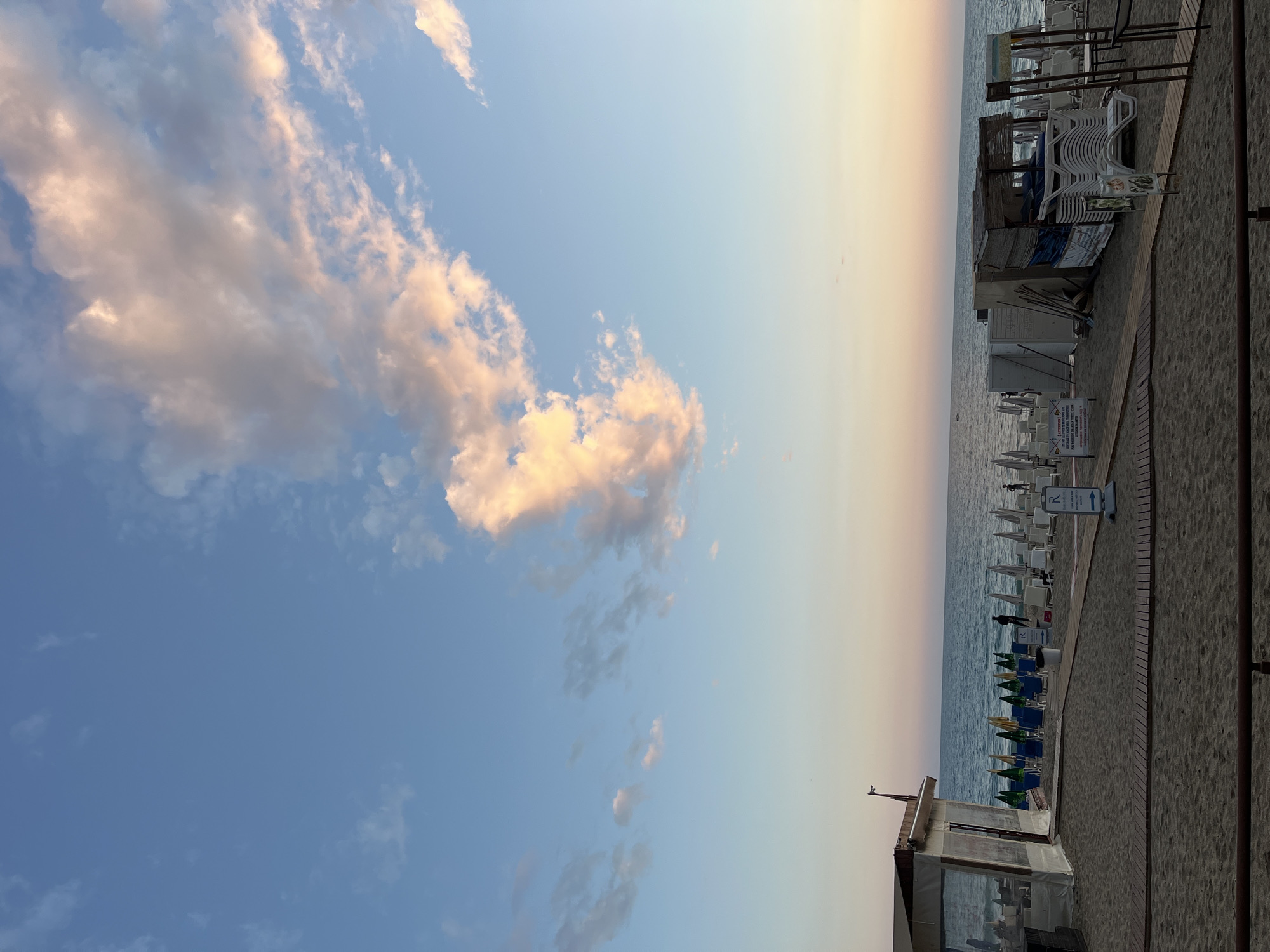 The image depicts a beach scene at sunset in Alanya, Turkey. To the left is a blue sky with light, puffy clouds tinged with orange. To the right is a beach with stacked white beach chairs, beach umbrellas, and a small building. There are a few people visible on the beach. The horizon line is pale orange. The image is taken vertically.