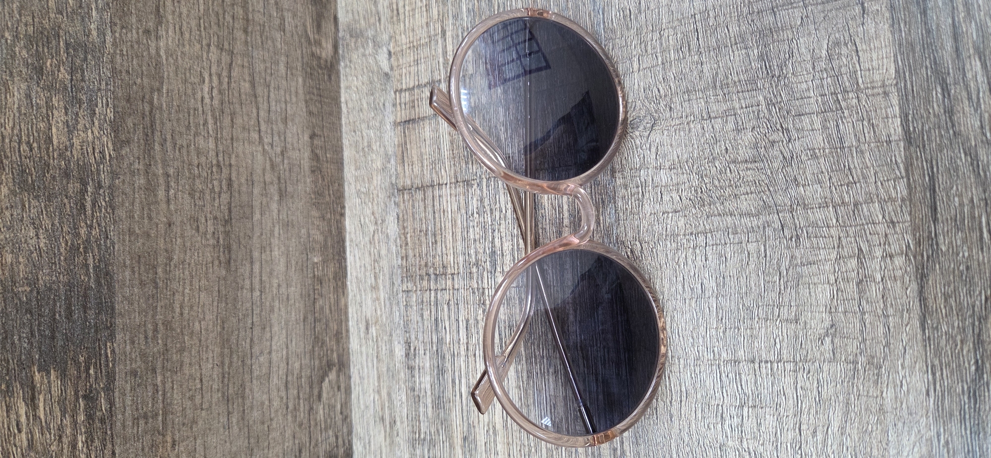 The main image displays a pair of contemporary sunglasses resting on a textured wooden surface. The sunglasses feature large, circular, dark lenses with subtle reflections, likely indicating an indoor light source or window. Their frames are a light, translucent material, possibly a soft pink, rose gold, or clear amber color, and the arms are folded inwards. The design has a modern or retro-inspired aesthetic.
The wooden surface, which appears to be a floor or tabletop, exhibits a prominent and varied grain pattern. Its coloration shifts from darker browns and grays on the left side to lighter, faded tones towards the right, suggesting a distressed or rustic finish. The wood grain runs predominantly vertically across the image, contributing to the overall texture of the background.
No individuals are present in the image, and no specific activity is taking place; it is a still life composition. There are no discernible cues regarding the time of day, weather, or a precise location beyond an implied indoor setting. No visible text is present within the image. The accompanying crops offer closer examinations of both the intricate wood grain and the detailed construction of the sunglasses' frames and lenses, highlighting their textures and colors.