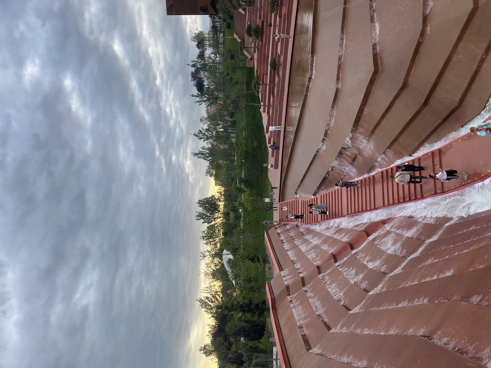 The image shows a tiered, cascading fountain in Krasnodar, Russia. The fountain is constructed of rust-colored stone or concrete. Water flows down the tiered structure. Several people are walking along the paths within the fountain complex. Green trees and lawn border the fountain. The sky is overcast with cloudy conditions.