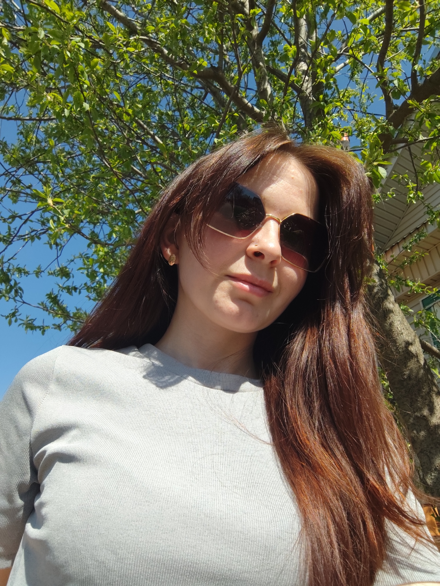 A low-angle, full-color photograph depicts a woman wearing sunglasses with a tree in the background and part of a building to the right. The woman has long, reddish-brown hair and is wearing a light gray ribbed shirt. The sunlight is shining on her face. The background is out of focus, but green leaves and blue sky are visible, as well as what appears to be part of a light-colored building. Based on the prompt, the image would have been taken in the city of Фоки, Russia.