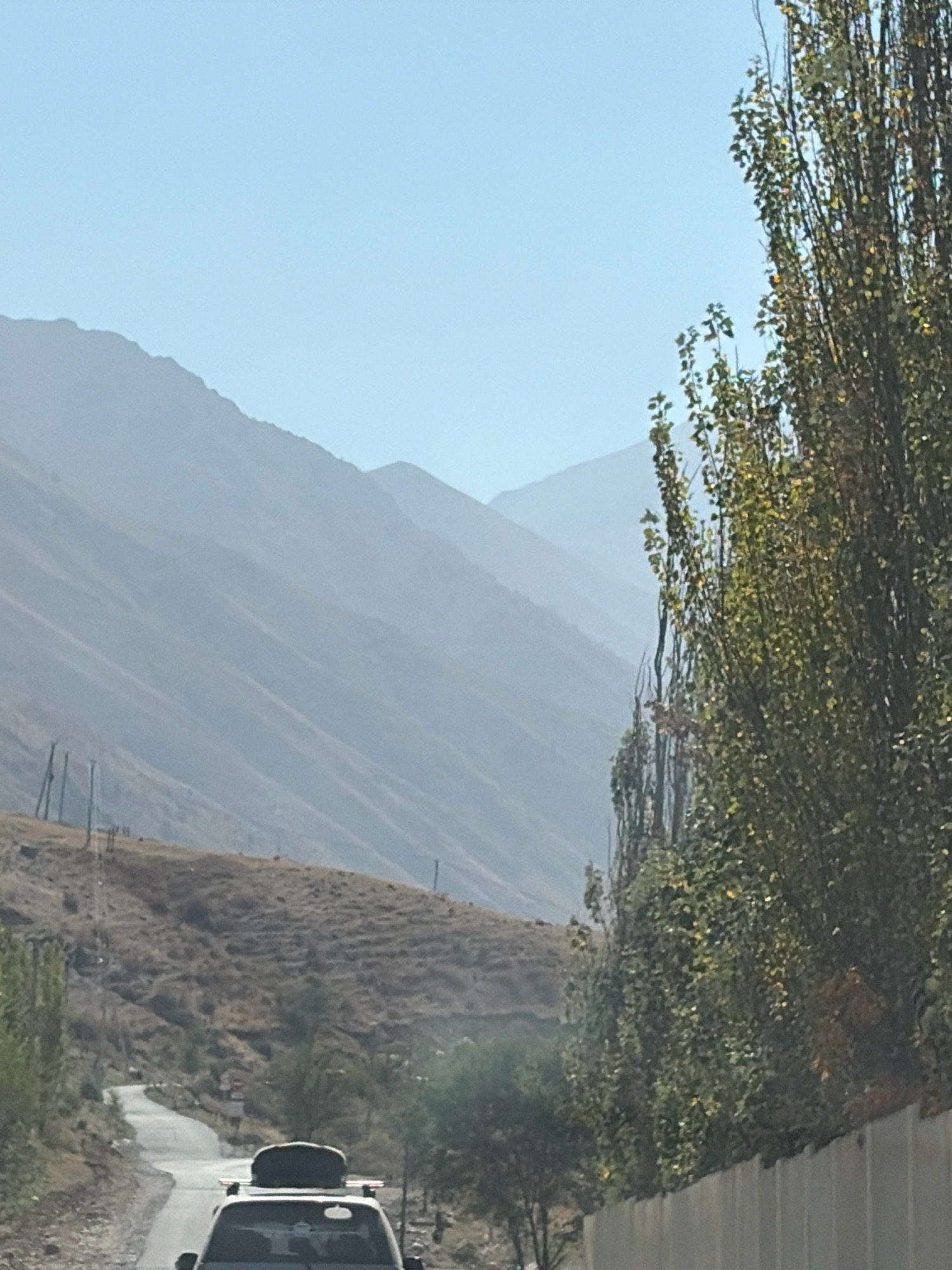 A car with luggage on its roof drives on a paved road that winds through a barren, mountainous landscape. The road is empty except for the car, which is the primary subject. The mountains in the background are hazy and layered, fading into the pale blue sky. To the right of the road, there are tall trees with sparse green leaves, and to the left, the landscape is covered in dry grass and sparse vegetation. The scene appears to be in a rural, mountainous region under a clear sky.