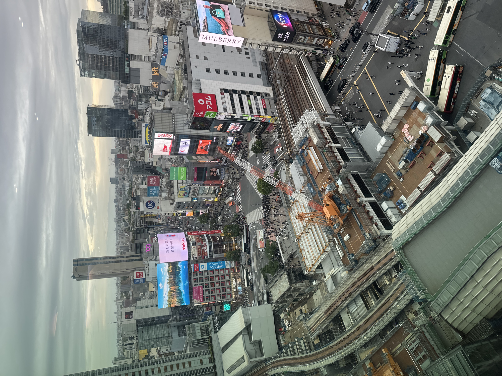 An aerial view captures the urban sprawl of Shibuya, Japan, distinguished by an amalgamation of skyscrapers and structures. The bustling area is punctuated by billboards advertising brands like Uniqlo and Yakult. Multiple trains, buses, and a multitude of people are seen navigating the streets and transit systems. Construction is underway in parts of the area, with a visible crane. The atmosphere is overcast, impacting the light and visual clarity of the scene.
