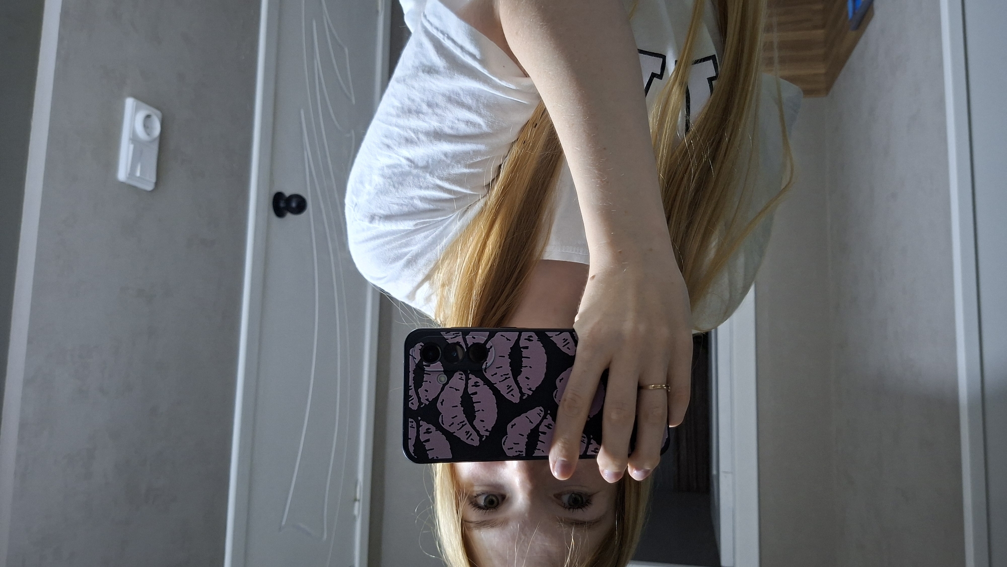 A person with blonde hair, wearing a white t-shirt and a ring on their finger, is holding a smartphone with a lip-patterned case to their face, obscuring it from view. The person is standing indoors, in front of a white door and a grey wall. The environment appears to be a room, possibly a bathroom or hallway, with a light switch visible on the wall. The image is taken from a medium shot, focusing on the person and their phone. The location is not determinable from the image.