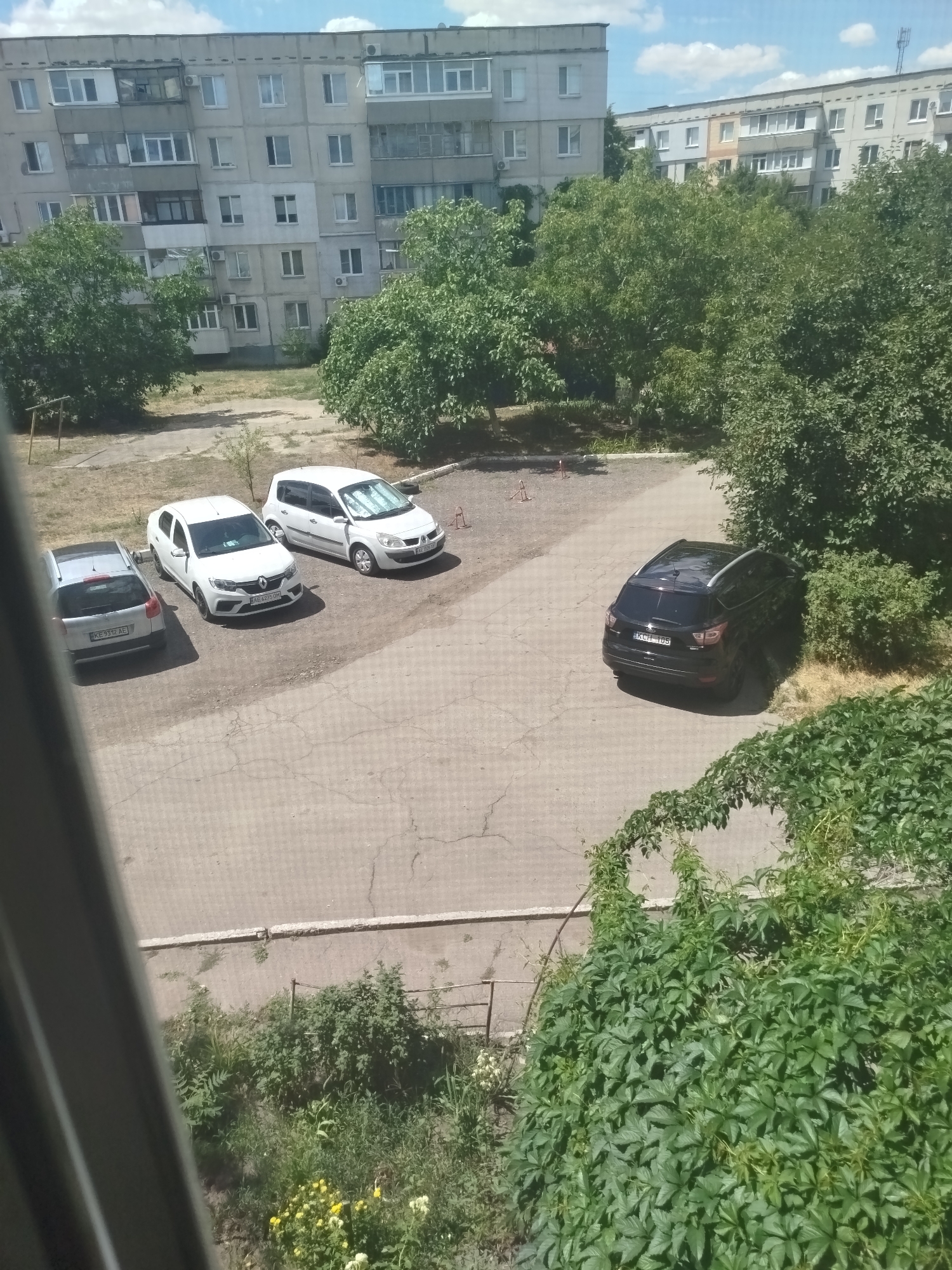 The image depicts a courtyard in a residential area, likely in Pavlohrad, Ukraine, characterized by apartment buildings, trees, and parked vehicles. In the foreground, a dense patch of green vines climbs a wall. Behind the vines, a paved courtyard area is visible, with several cars parked. To the left, a silver sedan is parked. Next to it, a white sedan, possibly a Renault Logan, is parked. Further to the right, a white hatchback, possibly a Renault Scenic, is parked. In the middle ground, a dark-colored SUV, possibly a Ford Escape, is parked. In the background, several multi-story apartment buildings dominate the skyline. Lush green trees are scattered throughout the scene, providing shade. The sky is partly cloudy. The overall impression is of a typical urban residential courtyard on a sunny day.