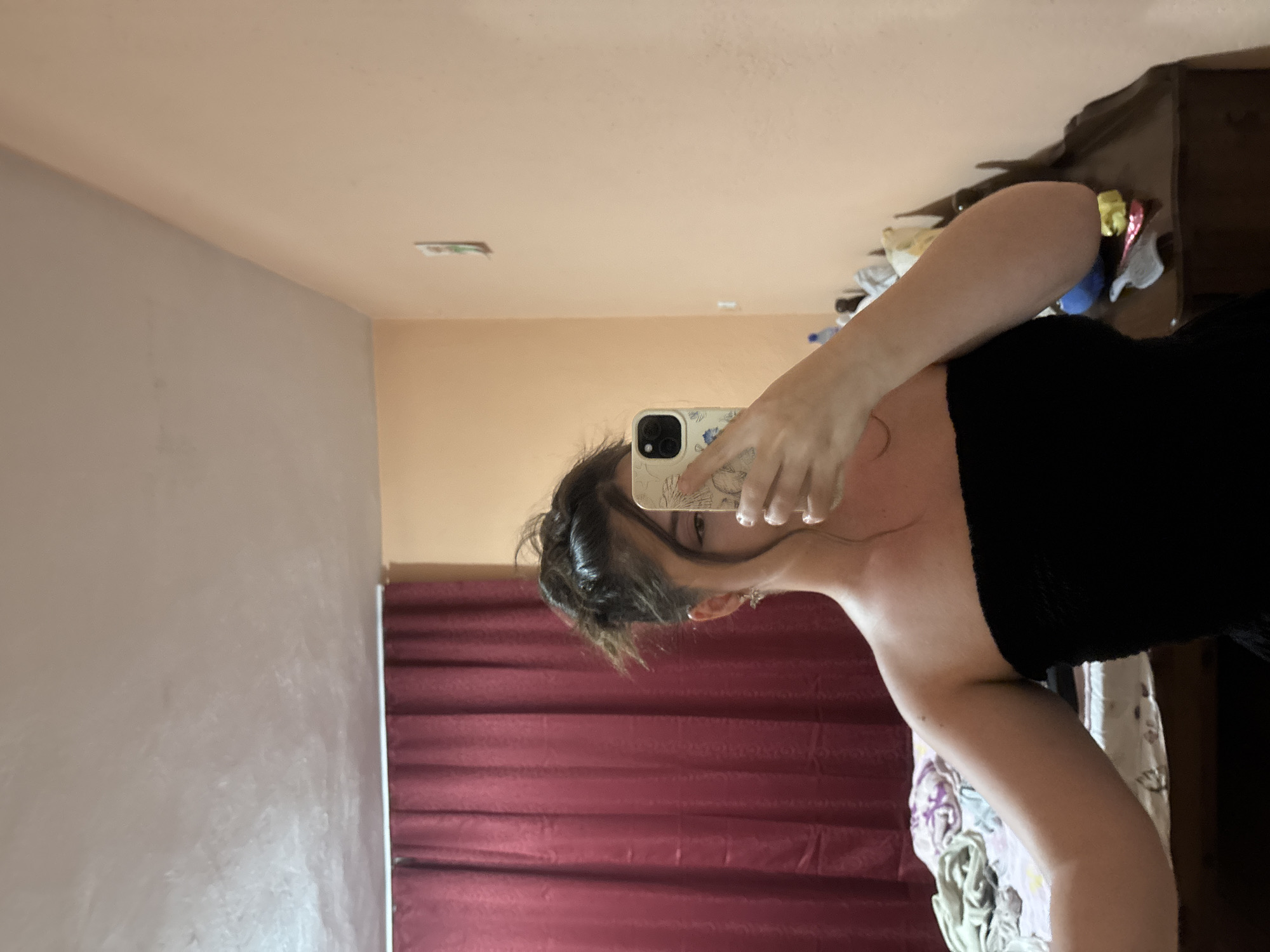 A young woman, partially obscured by her phone, stands in what appears to be a bedroom. She is taking a selfie in a mirror, wearing a black strapless top. Her hair is pulled up loosely. A dark red curtain hangs behind her to the left, and a wooden dresser is visible to the right. The walls are painted a light peach color, and the ceiling is a pale, textured white. The overall impression is of an informal personal space. The image does not contain enough information to determine its location in the city of Garmen, Bulgaria.