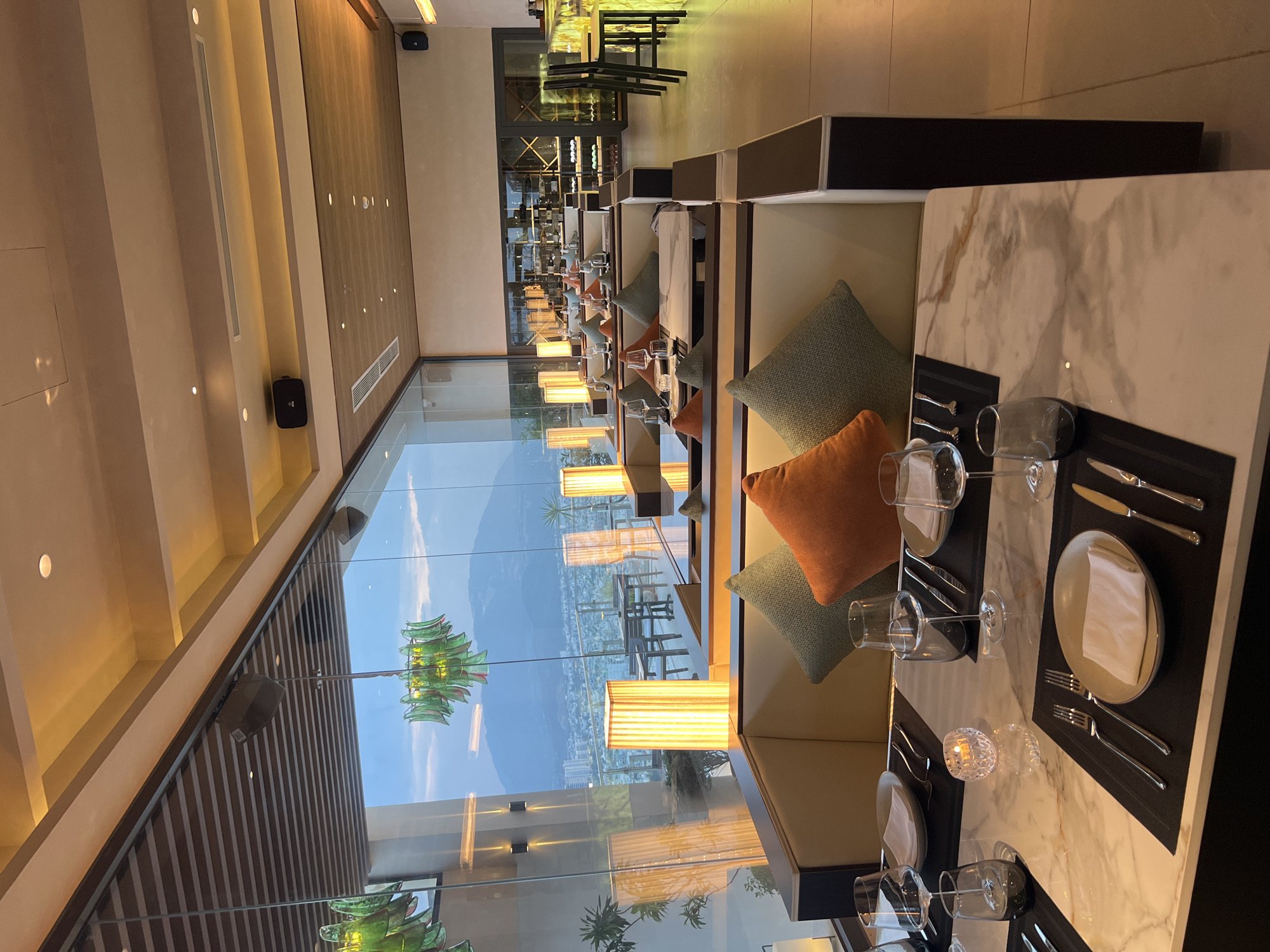 Here is a detailed description of the image:
The image captures an interior view of a high-end restaurant, potentially located in a city with a scenic mountain vista. The restaurant features floor-to-ceiling glass walls that offer a panoramic view of the mountainous landscape and a cityscape in the distance, suggesting a location elevated above the city.
The foreground focuses on the dining area, which is elegantly set up with tables ready for service. The tables have marble tops and are decorated with black place mats, plates, cutlery, and glassware. Some tables are adorned with decorative elements such as small, ornate candle holders. Each table is accompanied by a booth-style seating arrangement with plush cushions in varying shades of green and orange, indicating a comfortable and sophisticated dining atmosphere.
The interior lighting is warm and inviting, achieved through ambient fixtures and table lamps that create an intimate setting. The overall design of the restaurant seems modern and upscale, with attention to detail evident in the decor and layout.
In the background, a bar area is visible, stocked with a range of bottles, suggesting a selection of alcoholic beverages. The restaurant appears to be well-maintained and clean.
The environment suggests a fine dining experience in a location that leverages its elevated position to offer customers stunning views while they enjoy their meals.