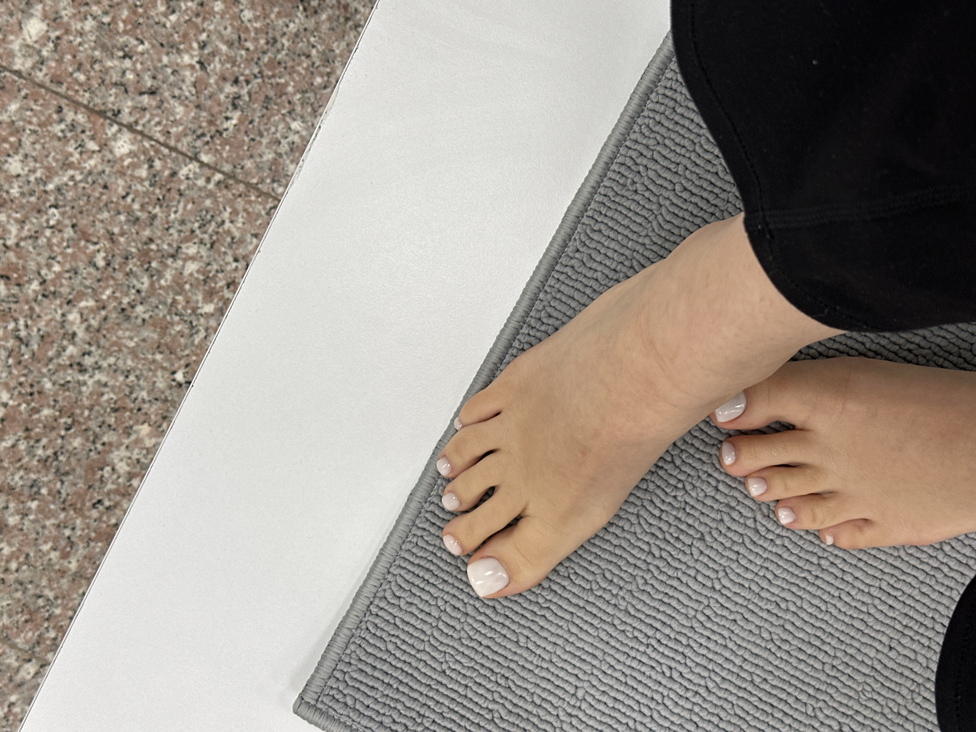 The image shows a close-up view of a person's feet resting on a grey mat. The person is wearing black pants. The toes have pale nail polish. The mat is placed next to a white surface and a speckled tile floor. The location in Sofia, Bulgaria, is not discernible from the image.