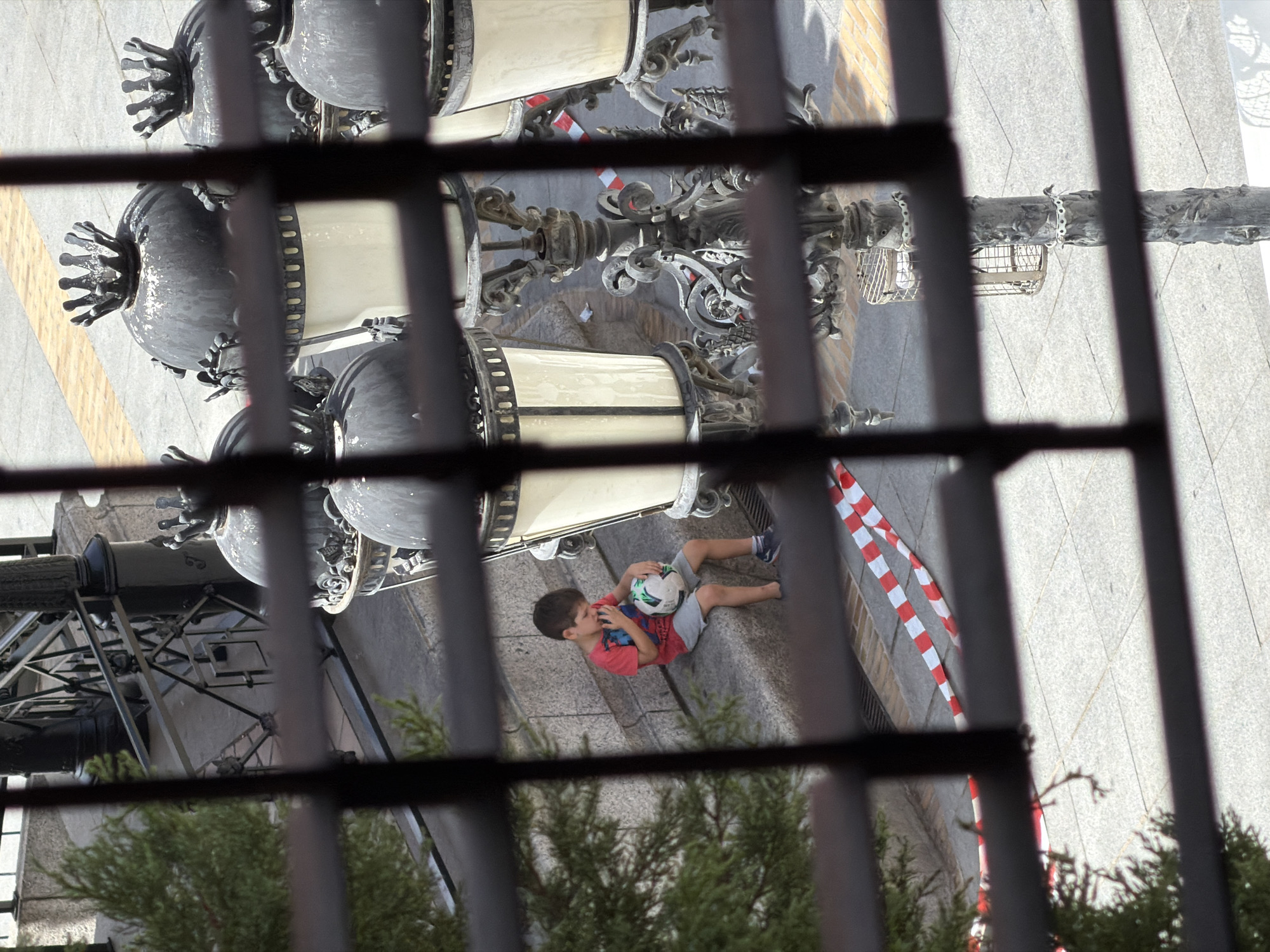An outdoor medium shot shows a young boy sitting on stone steps holding a soccer ball. The boy is wearing a red t-shirt and grey shorts and has his hand up to his mouth. He is looking down at the ball, which is white with green accents. Behind the boy, a street lamp with multiple lights is visible. In the foreground, a dark metal grate obscures the view. The area is paved with stone and there is red and white striped tape on the ground. The background includes a metal railing and a small green tree. The scene appears to be in a public area of Las Rozas de Madrid, Spain.