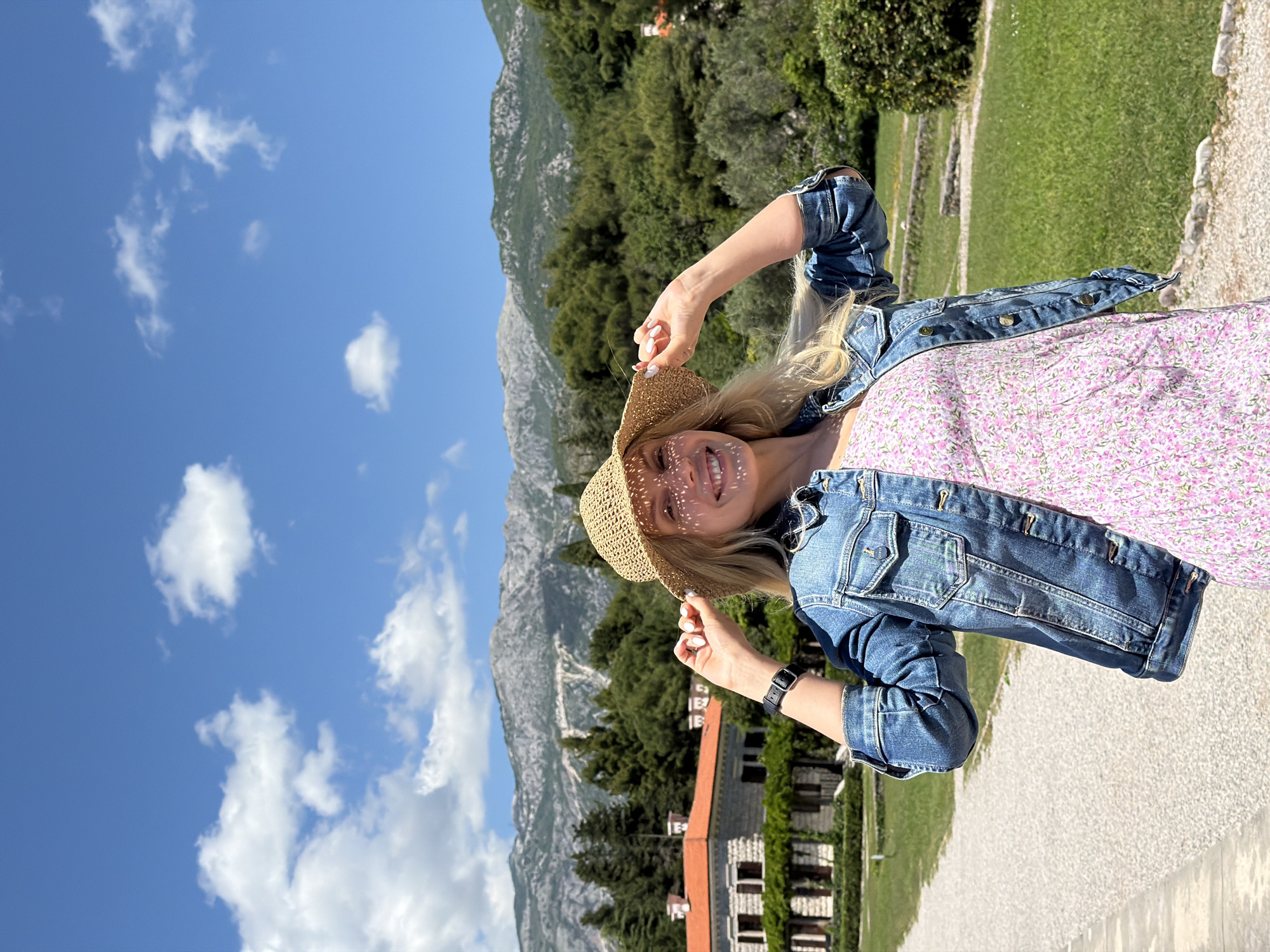 A woman in a straw hat and denim jacket stands on a gravel path with mountains and a building in the background. The sky is blue with scattered clouds. The woman is fair-skinned with blonde hair, and she is smiling. The building has a red tile roof and stone walls. The location is Sveti Stefan, Montenegro.