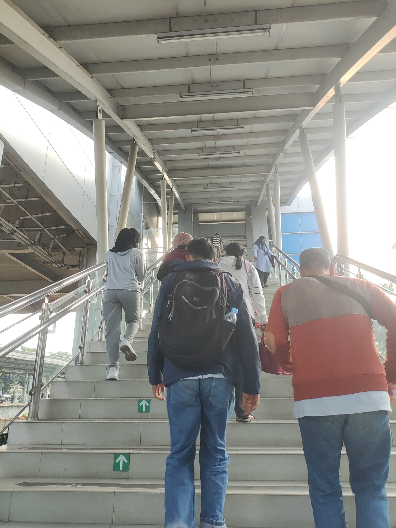 The image shows a group of people walking up a set of outdoor stairs under an elevated walkway. The walkway has a metal beam and concrete ceiling structure. The stairs are made of concrete and have metal handrails. Several people are visible from behind, dressed in casual clothing. Some are wearing backpacks. Green arrow signs pointing upwards are visible on some of the steps. The scene appears to be in a public transportation hub in East Jakarta, Indonesia, possibly a train station or an elevated bus stop, given the architectural style and the presence of commuters. The lighting suggests it is daytime, possibly late afternoon, with bright sunlight visible on the right side of the image.