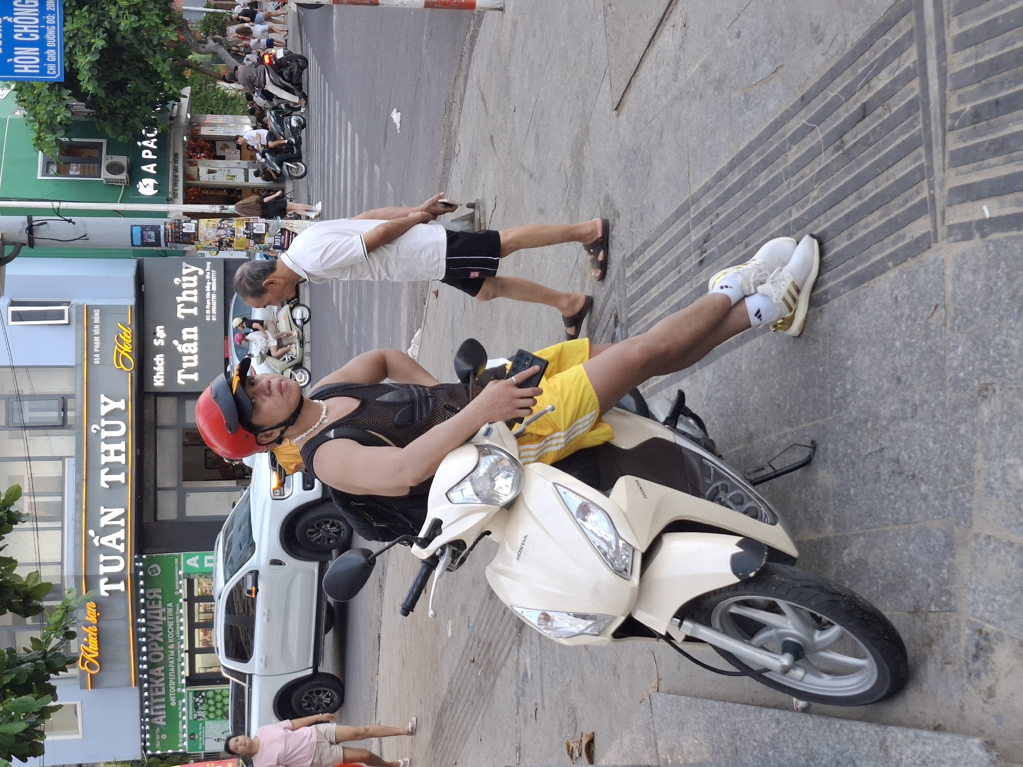 This media file depicts a street scene in Bac Nha Trang Ward, Vietnam. The primary subjects are several people and various objects, including motorbikes and a white pickup truck. The setting appears to be a commercial street with businesses, evident from signage.
In the foreground, a person is seated on a cream-colored Honda scooter, wearing a black mesh tank top, yellow shorts, a red helmet, and white athletic shoes with gold accents. They are holding a dark-colored object, possibly a phone, in their right hand. To their right, another person, wearing a white t-shirt and black shorts, stands next to a parked motorbike. Further in the background, other individuals can be seen walking or standing near buildings and vehicles. A white pickup truck is parked on the street.
Visible text includes "Khách sạn TUẤN THỦY", "05A PHẠM VĂN ĐỒNG", "Hotel", "АПТЕКА ОРХИДЕЯ", "ФИТОПРЕПАРАТЫ & КОСМЕТИКА", "A PÁC", and "HÒN CHỒNG". These suggest a hotel and a pharmacy are nearby. The "HÒN CHỒNG" text also indicates a specific location, possibly "Hon Chong" in Nha Trang.
The lighting suggests it is daytime, likely late afternoon or early evening, given the shadows and the overall brightness. The scene is lively, with people present on the street, implying regular daily activity. The overall mood seems neutral and observational, capturing a slice of everyday life.