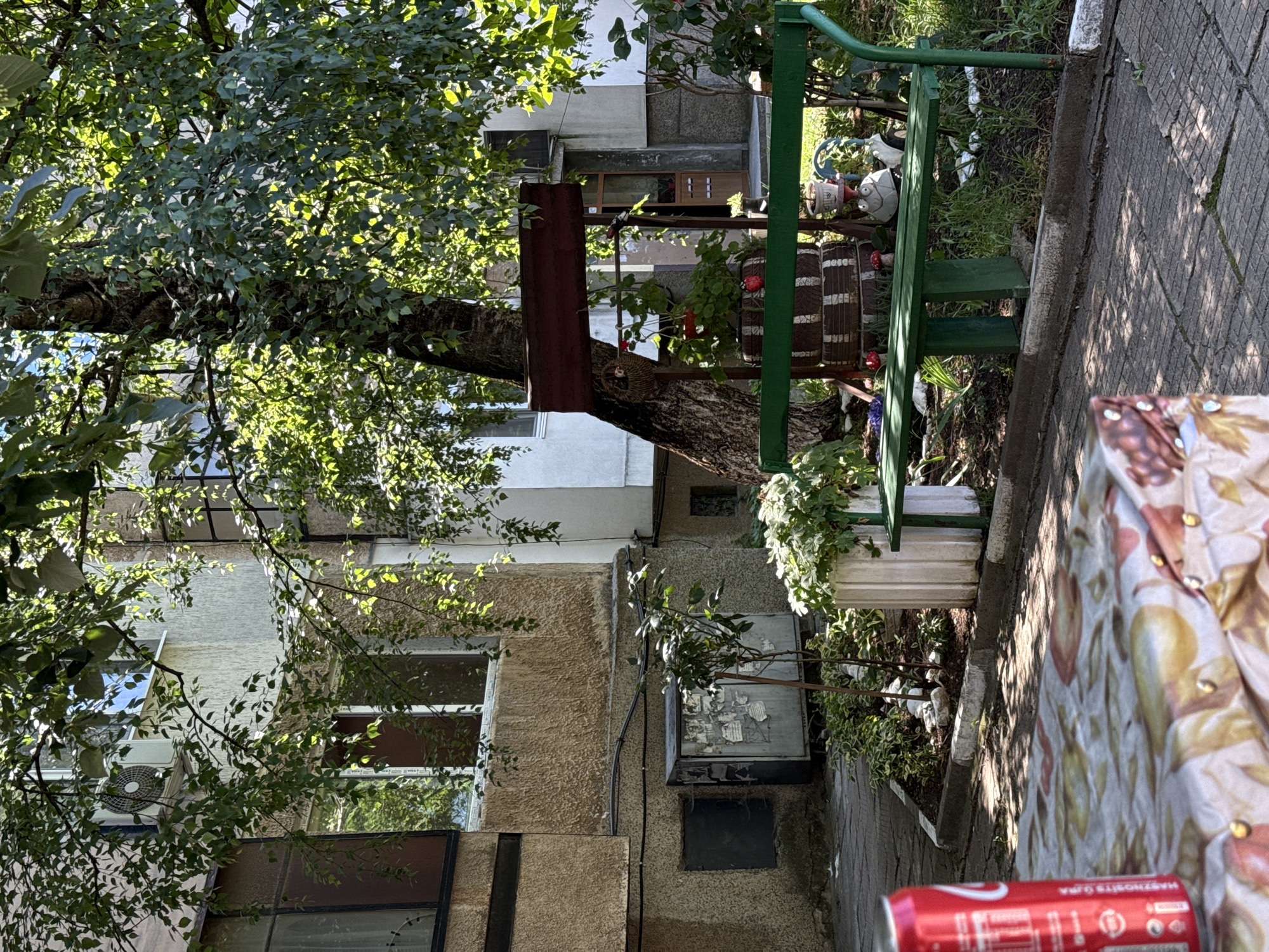 The image shows a sun-dappled courtyard in front of a residential building in Vratsa, Bulgaria. In the foreground, a table covered with a floral-patterned cloth is partially visible, with a red Coca-Cola can positioned on its left edge. Behind the table, a green metal bench is situated in a small garden area. The garden features a variety of plants, including a potted white plant and some smaller greenery, as well as decorative elements like stacked tires forming a rustic planter, ceramic mushroom figures, and a whimsical frog-like garden ornament. A mature tree with lush green foliage casts shadows over the scene. To the left of the garden, an outdoor air conditioning unit is mounted on the exterior wall of the building, which has a rough, textured finish and multiple windows. A metal utility box is also visible on the wall. In the background, the entrance to the building, with a wooden door, is slightly ajar, revealing a glimpse of the interior. The ground is paved with interlocking concrete tiles.