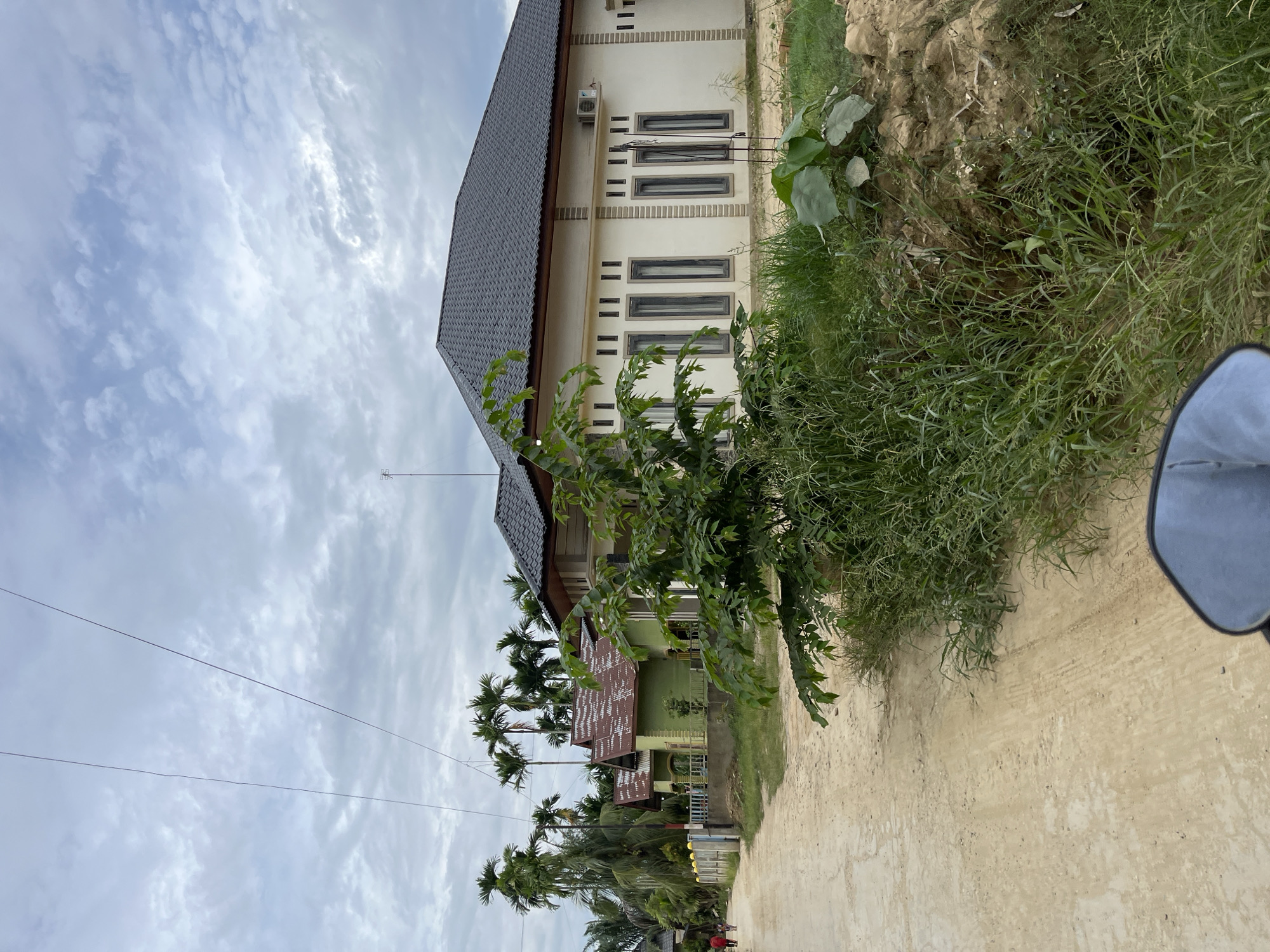 This image captures a scene in Menanggini, Indonesia, featuring a dirt road lined with houses and lush vegetation under a cloudy sky. On the right side of the road, a modern, single-story house with a dark tiled roof and cream-colored walls stands prominently. This house has several tall, narrow windows with dark frames and an air conditioning unit mounted on the exterior wall. In front of this house and to its left, dense green foliage, including tall grasses and a leafy tree, partially obstructs the view of the house.
Further down the road, to the left, are several other houses, some painted in lime green and others with reddish-brown tiled roofs. These houses appear to be of a more traditional style compared to the house on the right. A low fence, some sections with blue and white posts and railings, separates the properties from the road. Palm trees and other tall, slender trees with feathery fronds are visible behind these houses and along the roadside, contributing to the tropical environment. A few individuals, one wearing a red shirt, are walking on the dirt road in the distance. The road itself is unpaved, with tire tracks visible, suggesting it is frequently used by vehicles. The overall atmosphere is one of a residential area in a tropical, possibly rural or semi-urban, setting. The sky is overcast with thick, grey clouds. The perspective suggests the photo was taken from a low vantage point, possibly from a vehicle.