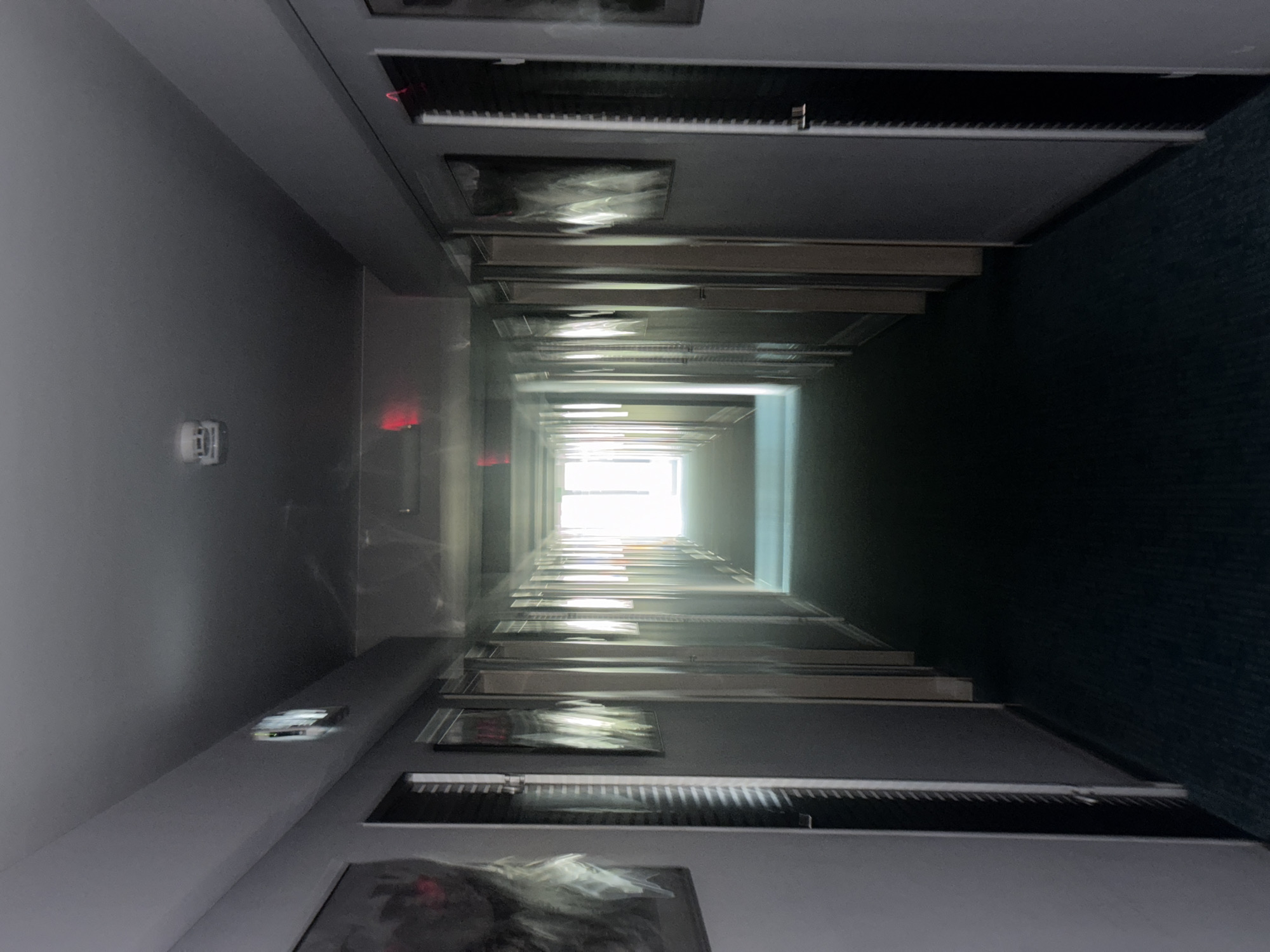 The image depicts a dimly lit, long hotel corridor with doors lining both sides. The corridor recedes into a bright, sunlit opening at the far end. Abstract framed artwork hangs on the walls, with some appearing slightly blurred due to motion or low light. A smoke detector is visible on the ceiling near the foreground. The floor is carpeted in a dark blue hue. The scene appears to be in an indoor, commercial establishment. There is no indication of the city of Yalta, Ukraine.