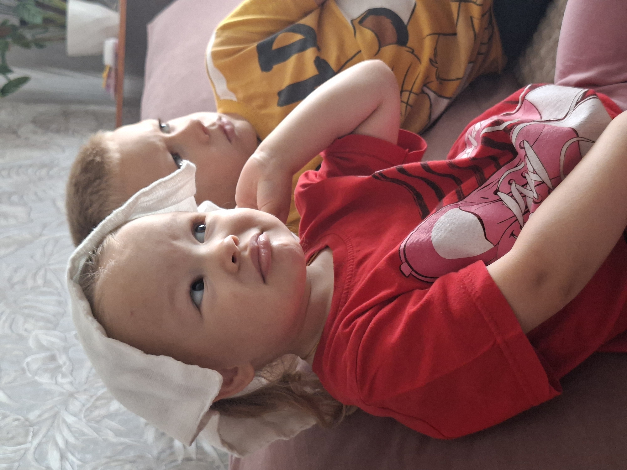 Two children, a girl and a boy, are sitting on a couch. The girl, in the foreground, is wearing a red t-shirt and has a white cloth draped over her head. She has light-colored hair and is looking to her right with a pouting expression. The boy, in the background, is wearing a yellow t-shirt with a graphic design and is also looking to his right with a serious expression. They are seated close together on a couch with a pink cushion. The background shows a textured wallpaper and a blurred potted plant on a shelf. The lighting suggests an indoor setting.