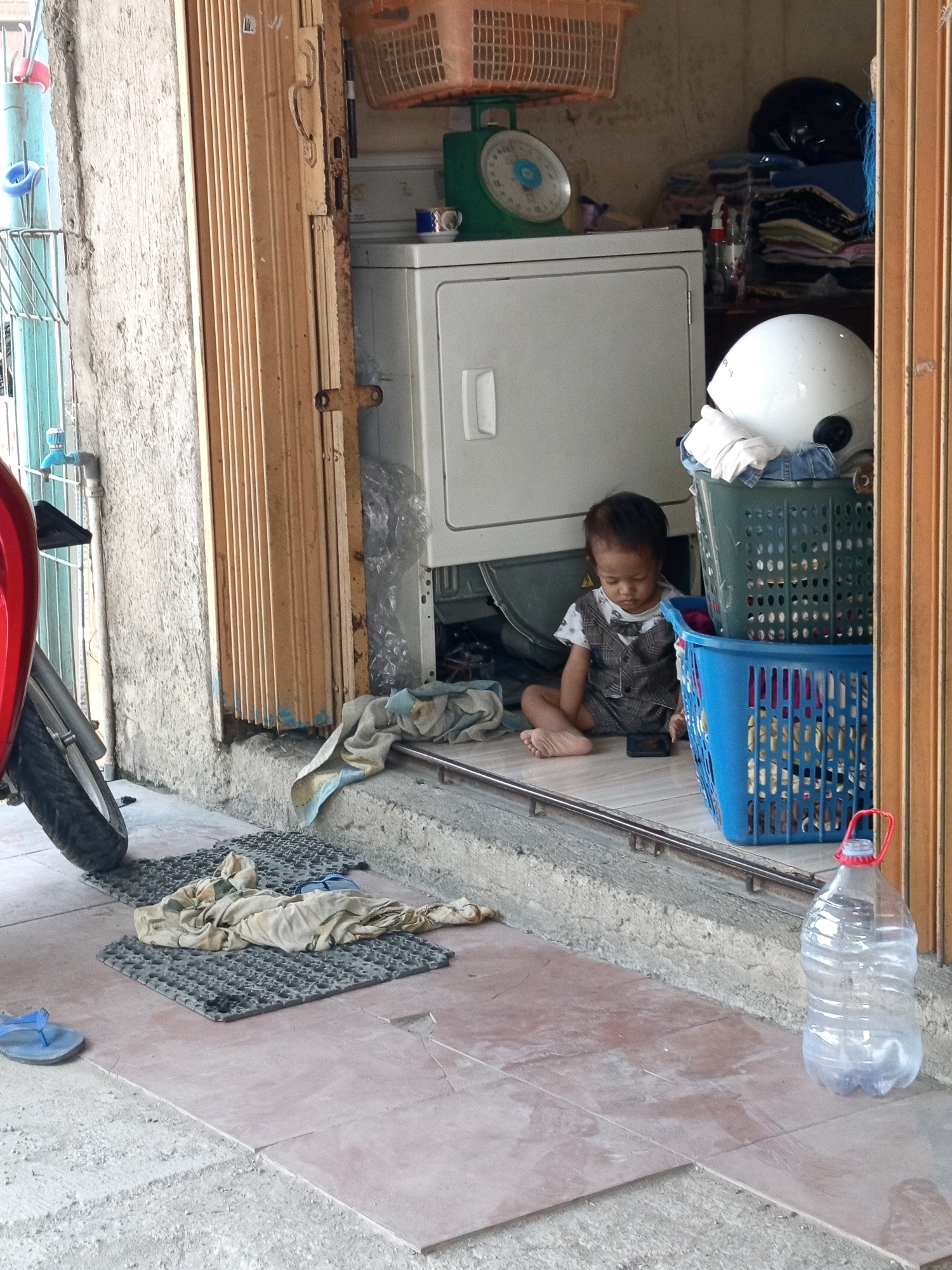 The image depicts an outdoor scene in Karawang, Indonesia. A young child, appearing to be a toddler, sits on the ground, looking at a mobile phone. The child is dressed in a patterned vest over a shirt. Next to the child are two plastic laundry baskets, one green and one blue, both filled with what appear to be clothes and other items. A white helmet rests atop the green basket, with some white fabric peeking out. In the background, a white washing machine is partially visible, along with a green scale topped with a wicker basket and a small cup on the scale. To the left, a red motorcycle is parked, with its front wheel and part of the body visible. Various items, including a blue flip-flop, a patterned cloth, and a black mat, are scattered on the ground in the foreground. A large, clear plastic bottle containing water is placed to the right of the laundry baskets. The ground is paved with reddish-brown tiles and concrete. The scene appears to be in front of a store or residence, with a corrugated metal door partially open on the right side of the frame.