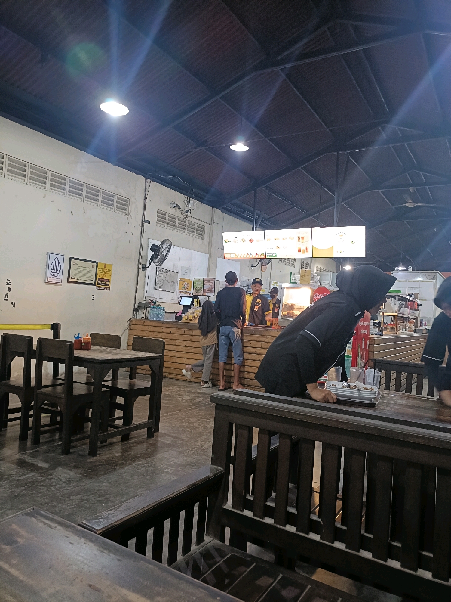 The image depicts the interior of a casual dining establishment, likely a food court or a restaurant with an open-air concept, located in Semarang, Indonesia. The scene is brightly lit by overhead fluorescent lights and ceiling fans are visible. The architecture features a corrugated metal roof supported by metal beams, and the walls are painted white.
In the foreground, several dark wooden tables and benches are arranged. One table in the left foreground has chairs and condiment bottles on it. A woman wearing a black hijab and black shirt is leaning over a table in the mid-ground, holding a metal tray.
Behind the tables, a counter area is visible where food is served. Several people are interacting at the counter, including staff members in uniforms and customers. A menu board with pictures of food items is displayed above the counter. Bottles of water are stacked near the counter.
A fan is mounted on the wall near the counter. Signs are posted on the wall, including one that appears to be a Halal certification from Indonesia. The floor is a concrete surface. The overall impression is that of a busy, informal dining space.