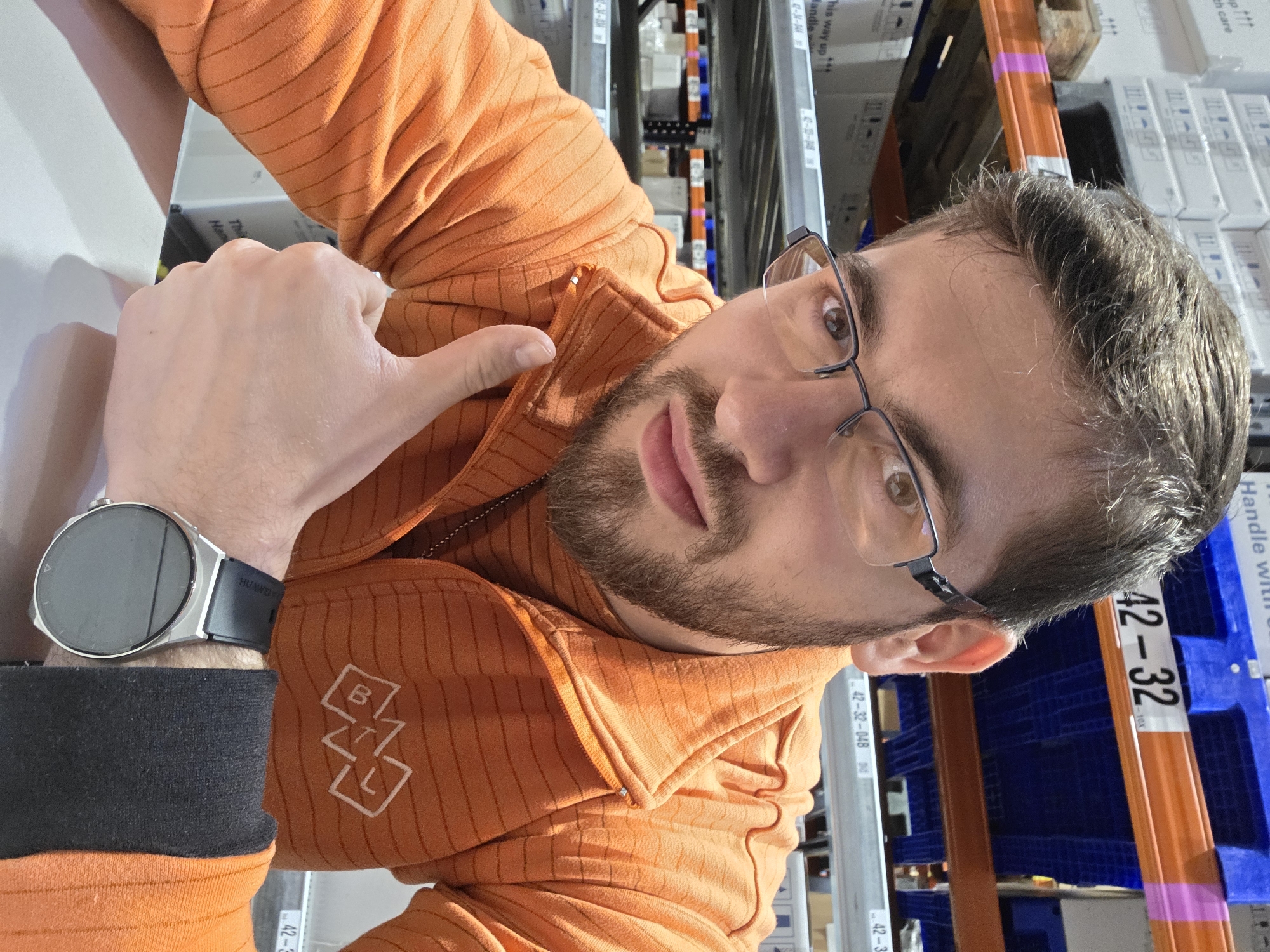 The image is a selfie of a man with brown hair and a beard, wearing glasses and an orange striped zip-up hoodie with a logo on the left side. He is wearing a smartwatch on his left wrist. He is giving a thumbs-up. The background appears to be a storage or warehouse setting with shelving units filled with boxes and containers. Label tags are visible on the shelves.