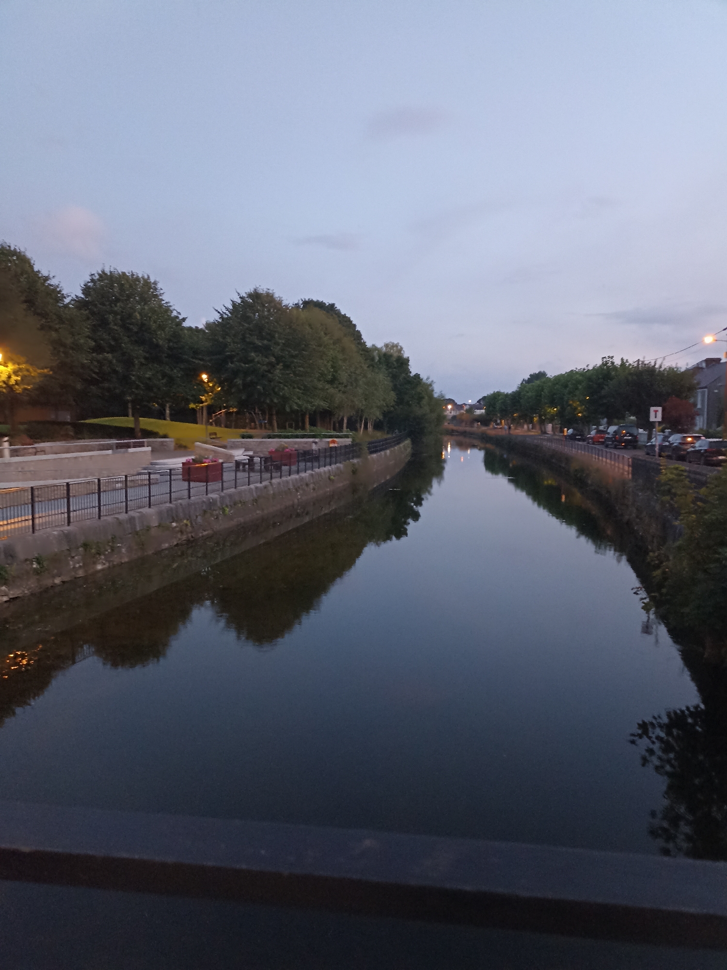The image depicts a tranquil canal in the city of Galway, Ireland, likely taken at dusk. The water surface reflects the twilight sky and the surrounding trees and buildings, creating a mirror-like effect. On the left bank, there is a paved walkway with a black metal fence, benches, and some greenery, suggesting a park or public area. Further along the canal, trees line the banks, interspersed with streetlights casting a warm glow. On the right bank, a street is visible with parked cars and buildings, indicating an urban setting. The overall atmosphere is peaceful and serene, characteristic of a canal scene in a European city.