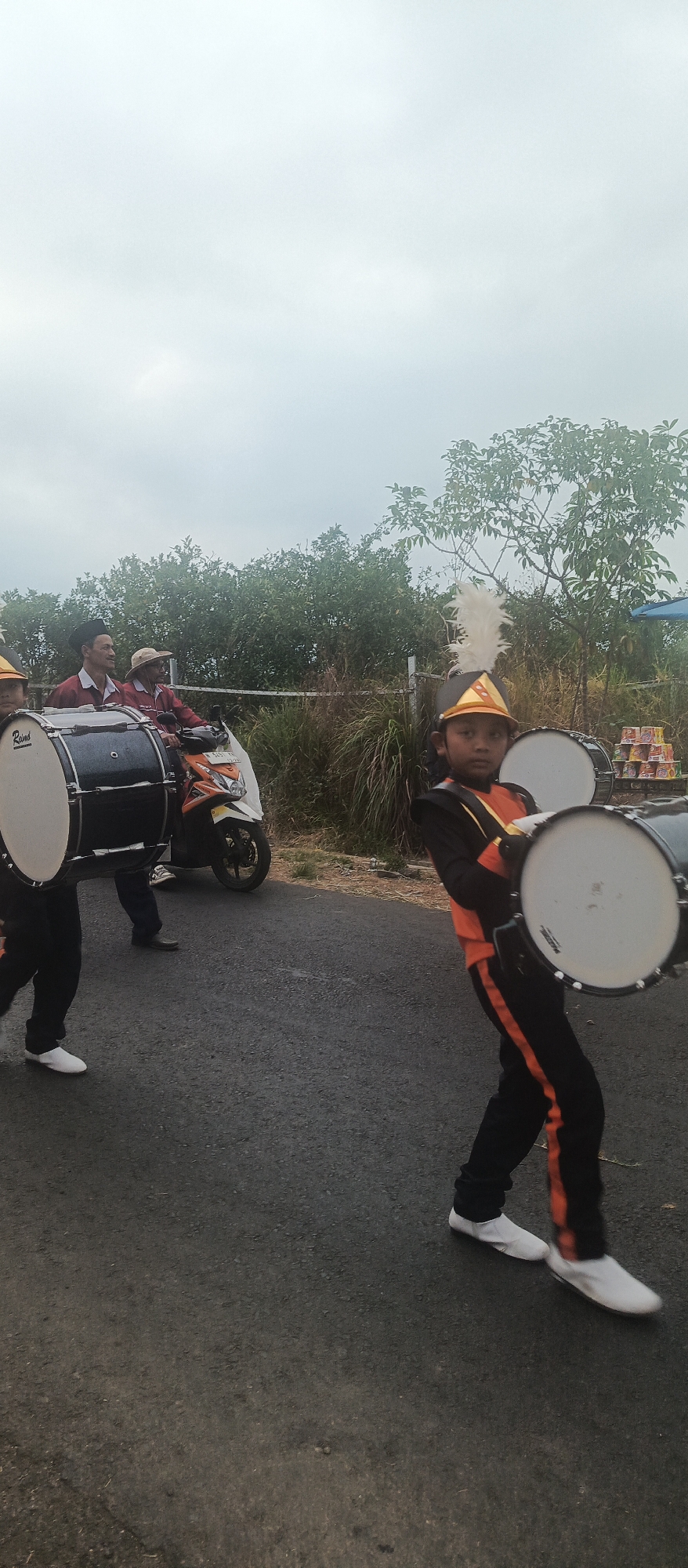This is a full-body, vertical, eye-level shot of a group of people, likely a marching band, on a paved road in Sumbersekar, Indonesia. The sky is overcast and pale blue. In the foreground, a young boy in a marching band uniform, with an orange and black jacket and pants, and a feathered headdress, holds two drums. To his left, another person, partially visible, also holds a drum. Behind them, two men are on a scooter, with one man in a red shirt and dark pants, and the other in a straw hat and a red shirt. The background consists of green trees and bushes, with a white fence. There are also some stacks of goods on a pallet in the background, to the right of the boy. The road appears to be asphalt. The overall scene suggests a parade or performance is taking place.