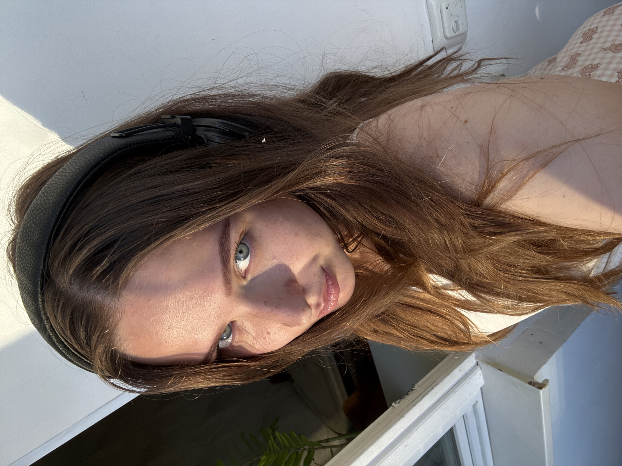 A young woman with long, brown hair and blue eyes is looking up at the camera, wearing black headphones. She is wearing a white top and has freckles on her nose and cheeks. She is positioned near a window with a white frame, and there is a plant visible in the background. The lighting suggests it is daytime, with sunlight casting shadows on the wall behind her. The image does not contain any information about the city of Chaikovsky, Russia.