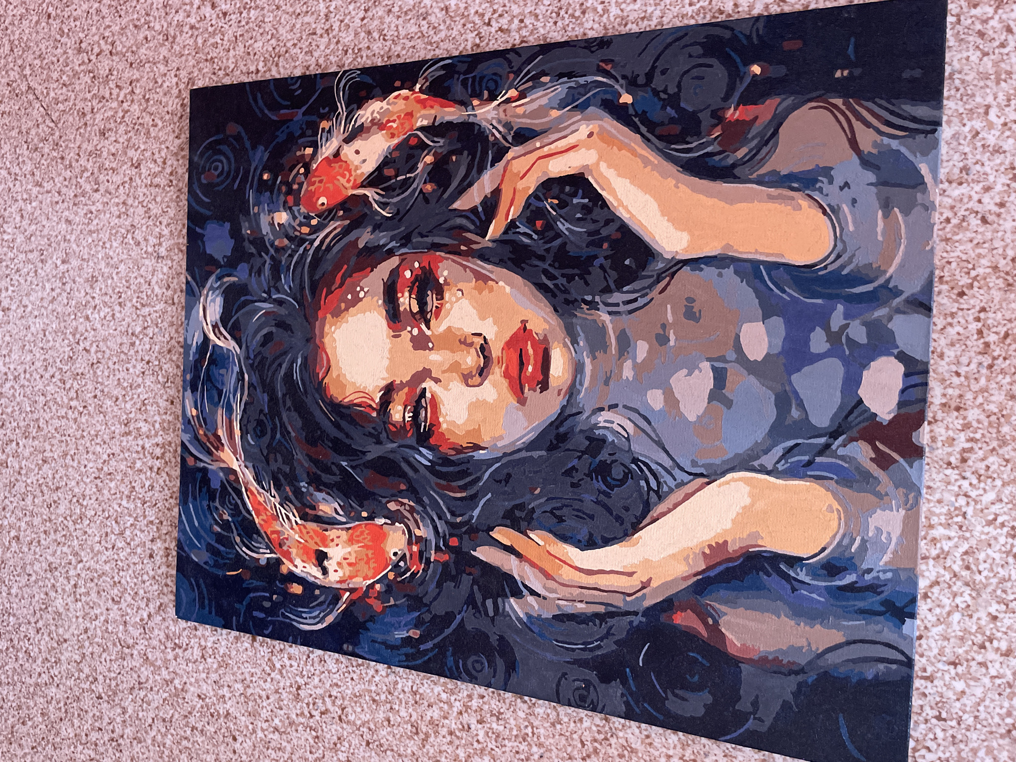 The image displays a close-up, high-angle shot of a painting resting on a textured, light-brown carpet. The painting itself is a portrait of a woman with dark, wavy hair, characterized by bold brushstrokes and a palette of dark blues, grays, oranges, and flesh tones. Two orange koi fish are depicted near her head, appearing to swim in her hair or surrounding water. The woman has striking red lips and eyes with prominent red eyeshadow. Her hands are raised towards her face, with fingers slightly splayed. The background is dark and abstract, with swirling patterns in shades of dark blue and gray. The painting appears to be a "paint-by-numbers" artwork, given the distinct sections of color and visible brushwork. The overall impression is one of a surreal or mystical depiction. The image does not contain any elements that would allow for the identification of a location in Laishevo, Russia.