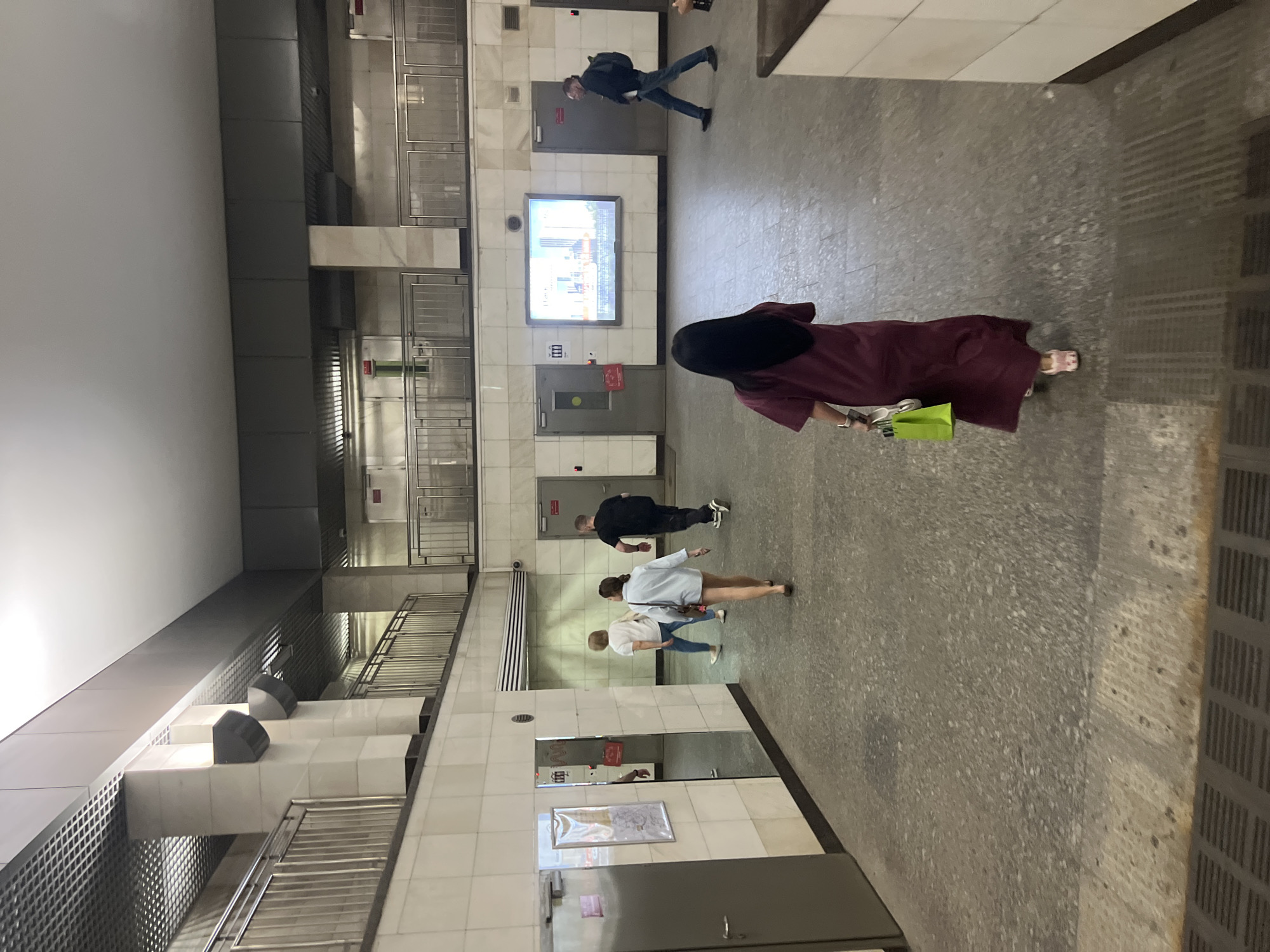 The image depicts an indoor public space, likely a metro station or subway concourse, with a concrete floor and tiled walls. In the foreground, a person wearing a long, burgundy dress walks away from the camera, carrying a bright green shopping bag. To their left, a large mirror reflects a person in a white shirt and blue jeans. Further into the scene, three people are walking away from the camera. Two are women in white tops and jeans, and one is a man in a black t-shirt and dark pants. In the background, there are several doors, a large illuminated advertisement, and a man in a dark jacket with a backpack walking towards the right. The overall environment appears to be utilitarian and modern, with metallic railings and suspended lighting fixtures. The location is identified as Kommunarka, Russia.