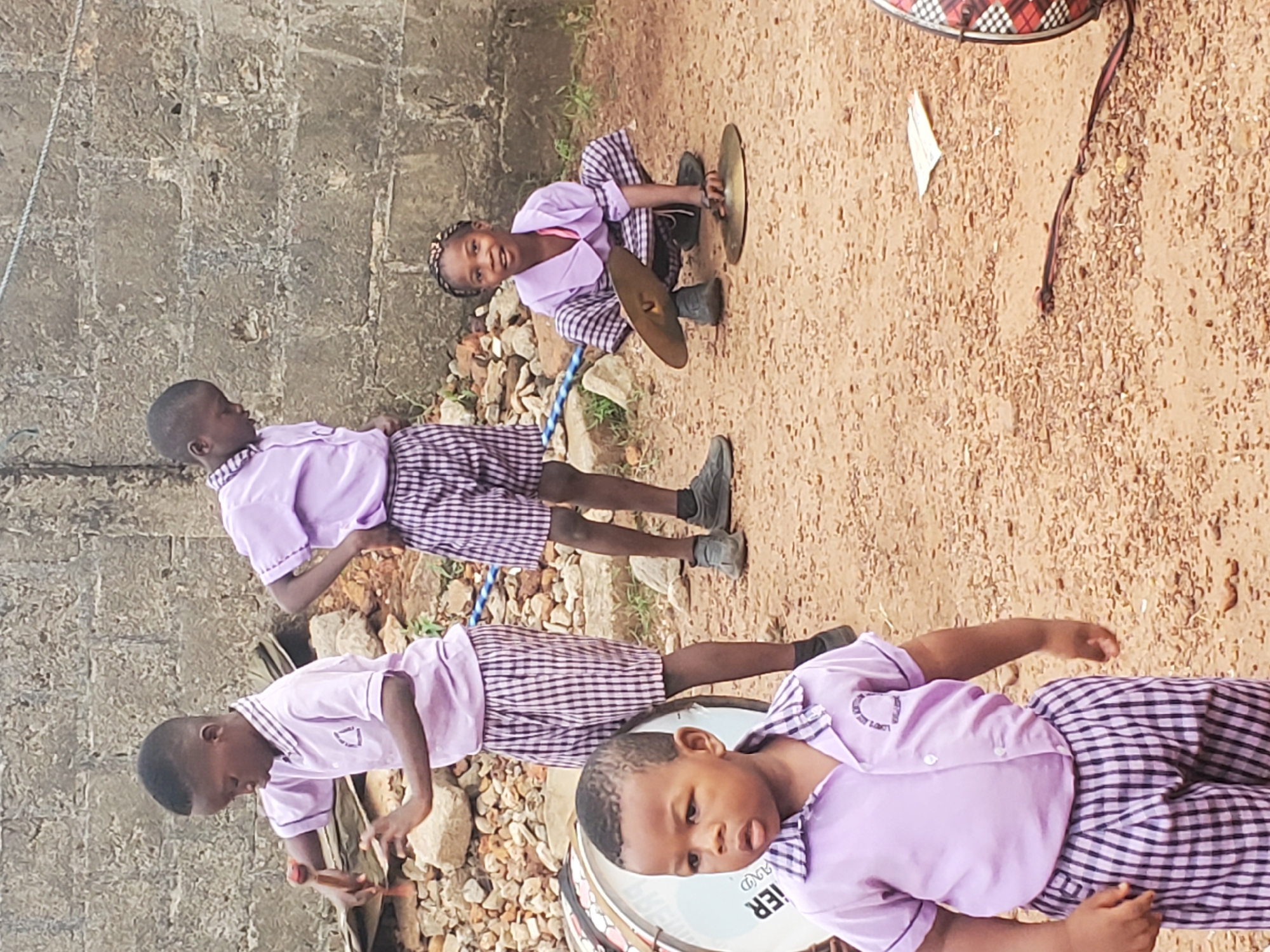 In Ukwu-Abwa, Nigeria, a group of four school children are outdoors on a dirt ground next to a brick wall. They are wearing light purple shirts and checkered shorts. One child is sitting and holding two cymbals. Another child is holding drumsticks and standing next to a pile of rocks. A third child is also standing with drumsticks. A fourth child is standing next to a drum.