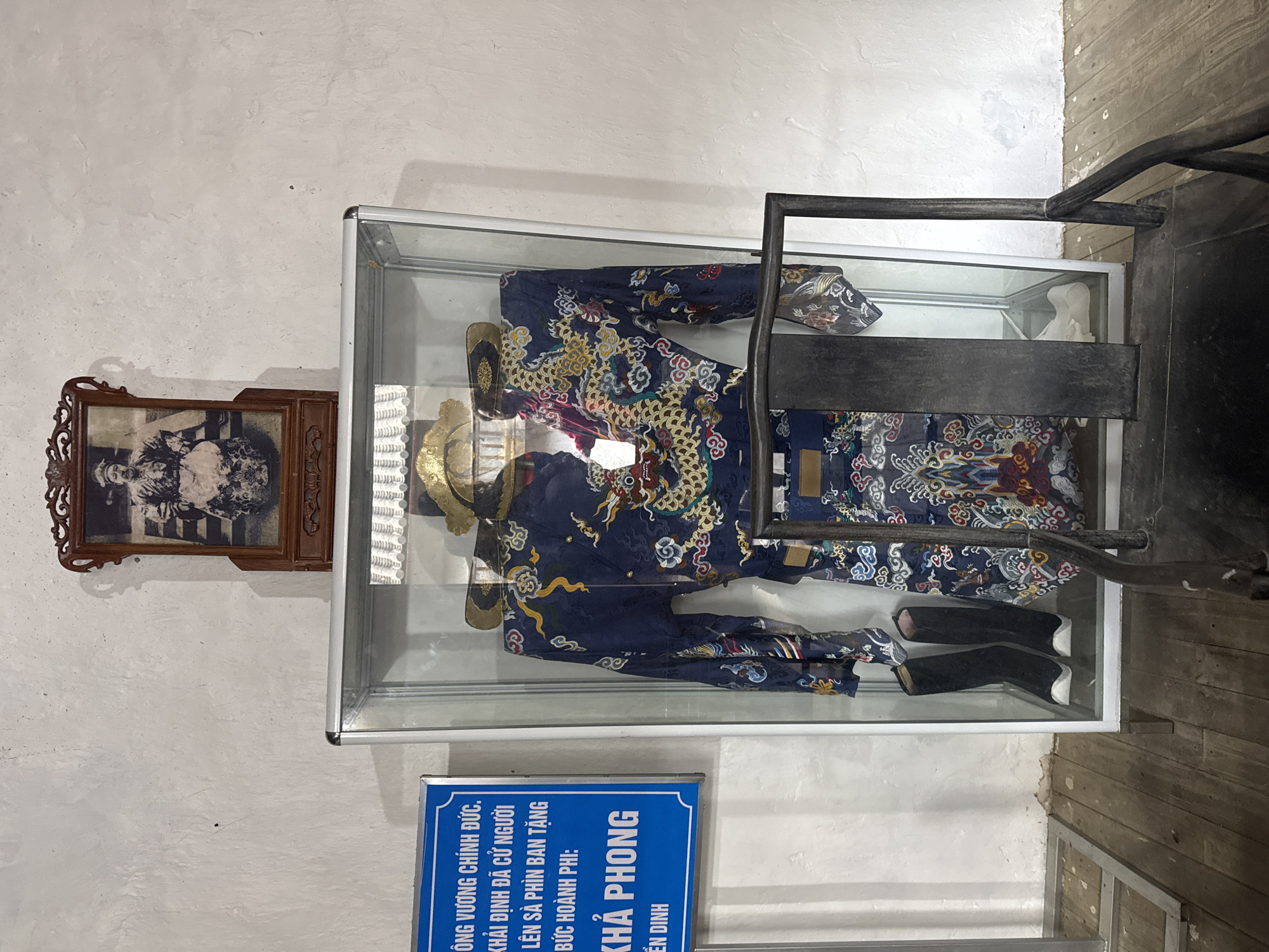 The image shows a display case mounted on a wall, containing historical artifacts. Inside the case, there is a traditional royal costume in dark blue with intricate gold and red patterns. Adjacent to the clothing are a pair of black shoes and a gold headpiece or crown. Mounted on the wall next to the display case is a framed photograph. Below the case, a blue informational sign with white text is visible. The Vietnamese text on the sign identifies the objects as being related to the Khải Định era and mentions Sà Phìn Commune, a location in Vietnam. The scene appears to be a historical exhibit in a museum or heritage site. The wall has a textured, painted surface. A portion of what looks like a metal structure is on the right side of the display.