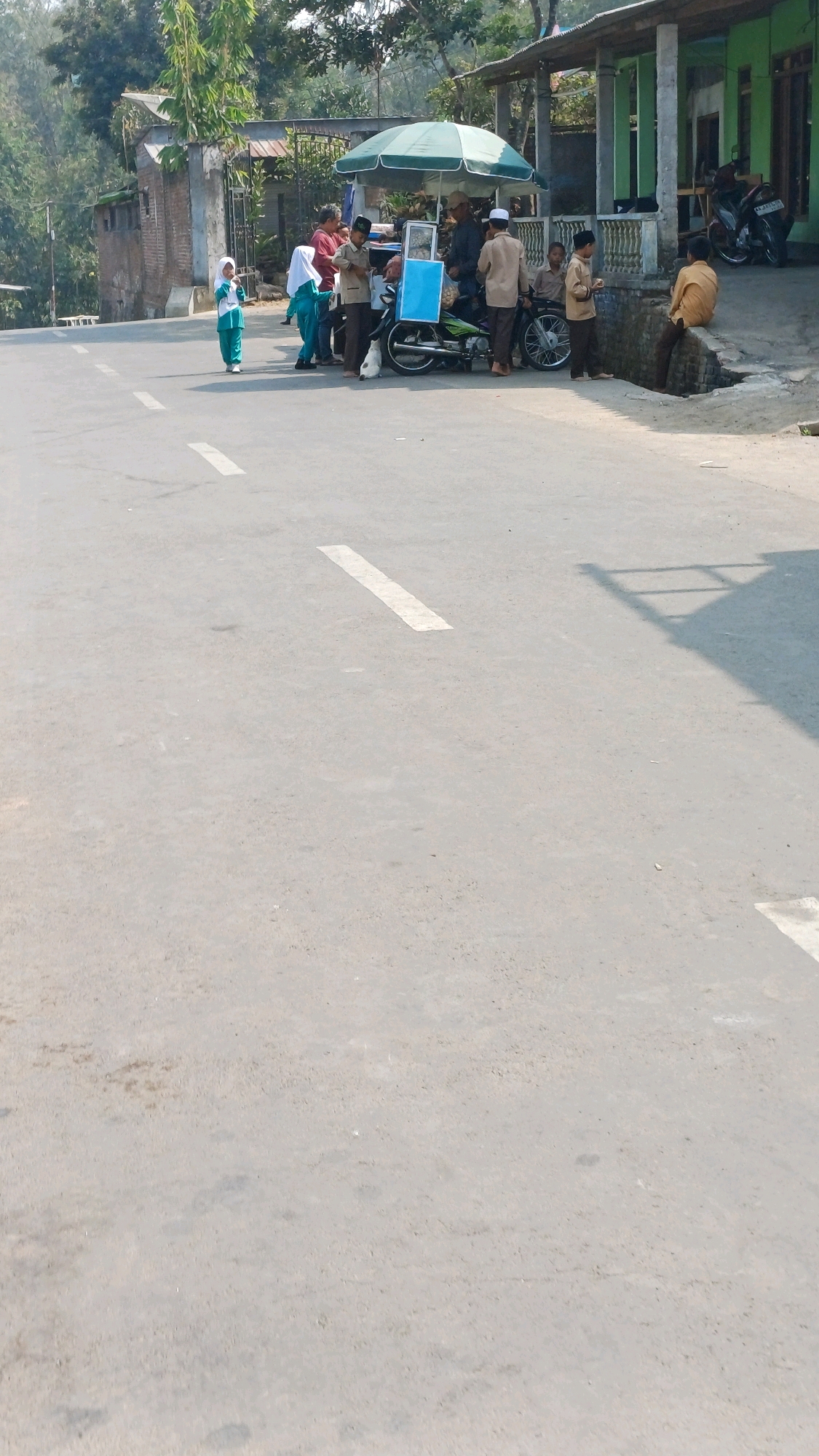 The image captures a street scene in Kupen, Indonesia, where a group of people are gathered around a food cart and motorcycles. In the foreground, the asphalt road features white lane markings. On the right side of the road, a small building with a green facade and pillars is visible. Adjacent to the building, a group of individuals, including children in what appear to be school uniforms, are interacting near a food cart covered by a green umbrella. Two motorcycles are parked nearby, one of which is fitted with a sidecar that displays a blue sign. Some adults are standing and conversing, while a few children are seated or standing. The general environment suggests a sunny day in a semi-urban or rural setting.