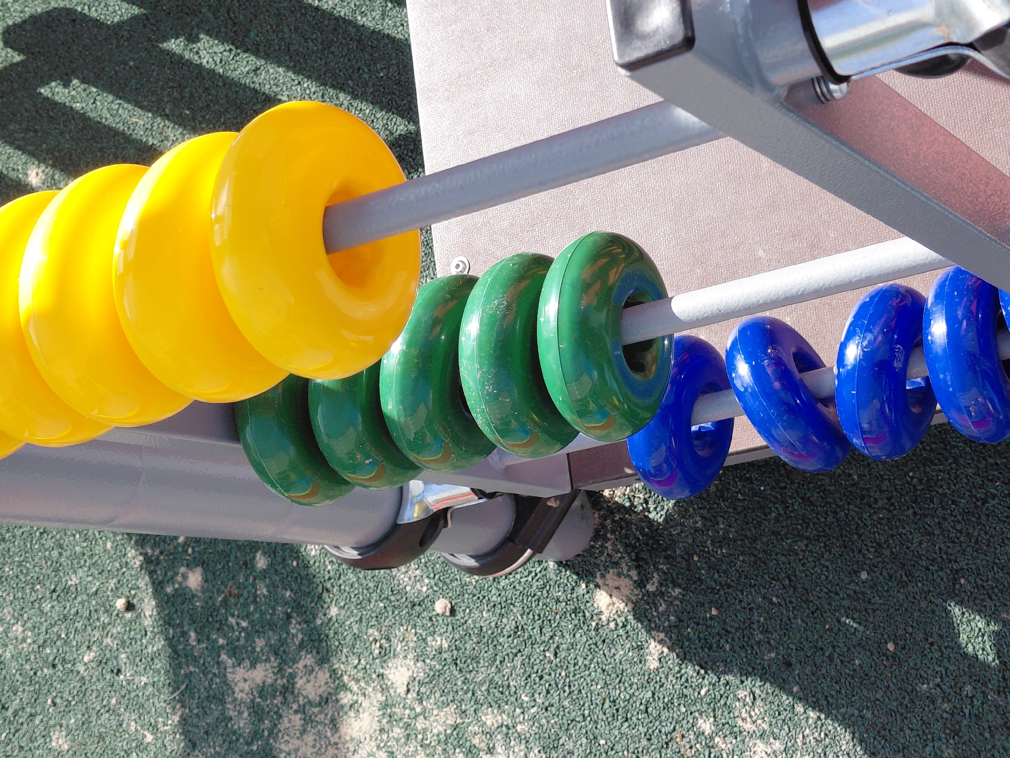 The image provides a close-up, high-angle view of a colorful abacus, which is part of a playground structure, situated outdoors in Nadym, Russia. The scene is brightly lit by natural daylight, casting distinct shadows on the ground.
The primary subjects are three rows of large, glossy plastic rings. The top visible row consists of five bright yellow rings, the middle row features five emerald green rings, and the bottom row displays four royal blue rings. These rings are threaded onto two horizontal metal rods: a grey rod for the yellow rings and a white rod for the green and blue rings. A robust grey metal frame supports these rods and the entire abacus assembly.
The ground beneath the equipment is a dark green, textured rubber safety surface, commonly found in playgrounds. Scattered light-colored specks, possibly sand or small debris, are visible on this surface. In the background, a light-colored, textured panel is partially visible, likely another component of the playground structure. The strong shadows suggest a sunny day, possibly mid-morning or late afternoon. There are no people visible, nor any discernible text in the image.