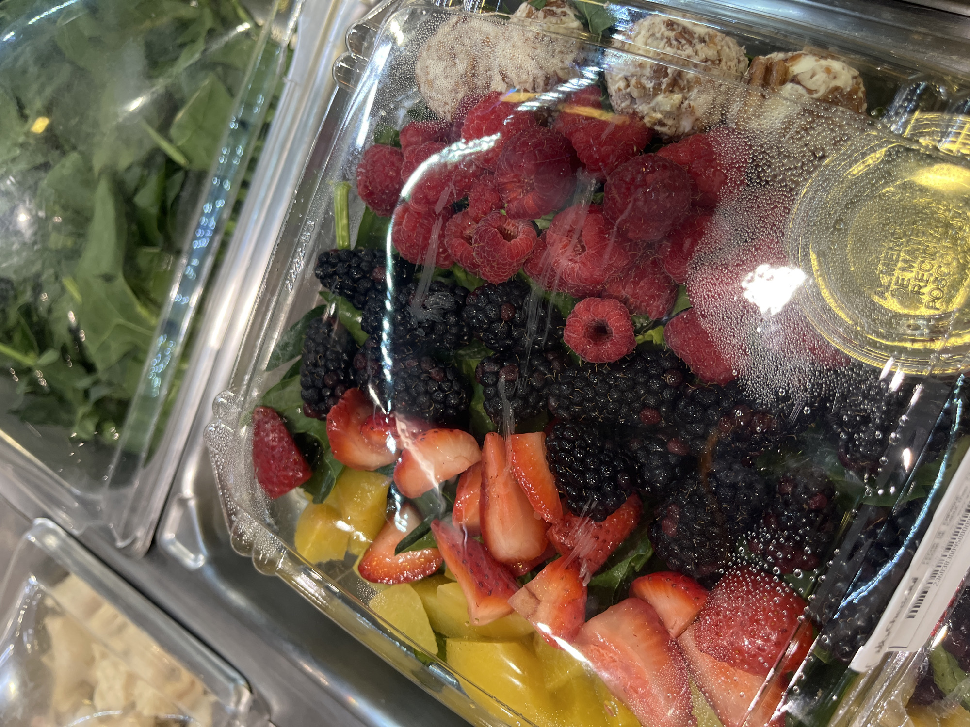 The image captures a close-up view of a pre-packaged salad in a clear plastic container, likely displayed for sale in a refrigerated section of a grocery store or deli in San Bernardino Tlaxcalancingo, Mexico.
The main subject is a vibrant fruit and cheese salad, carefully arranged over a bed of leafy greens, possibly spinach or mixed greens. The salad features a generous quantity of fresh berries, including bright red raspberries and dark, plump blackberries. Sliced strawberries are also visible, interspersed with diced pieces of a yellow fruit, which could be mango or pineapple. At the top of the container, there are two distinct, beige-colored, roundish items that appear to be some type of cheese, potentially goat cheese or a nut-crusted cheese.
The clear plastic lid of the container shows significant condensation and numerous small water droplets on the inside, indicating that the product is chilled or has been recently moved from a colder environment. A small, round plastic cup containing a clear, yellowish liquid, presumably salad dressing, is placed within the main container. On this cup, Spanish text is clearly visible: "AL MENOS DE MATERIAL RECICLADO POSIBLE," which translates to "AT LEAST FROM RECYCLED MATERIAL POSSIBLE," highlighting an environmental initiative.
A white label or sticker is partially visible inside the container, showing part of a barcode and a series of numbers: "2-2360300050000." Faint text, possibly "Mr. Chef," is also discernible on a label. The salad container rests on a metallic surface, with other similar clear plastic containers, including one filled with leafy greens, partially visible in the blurred background, reinforcing the setting as a commercial food display. The photo is taken from a slightly downward, angled perspective.