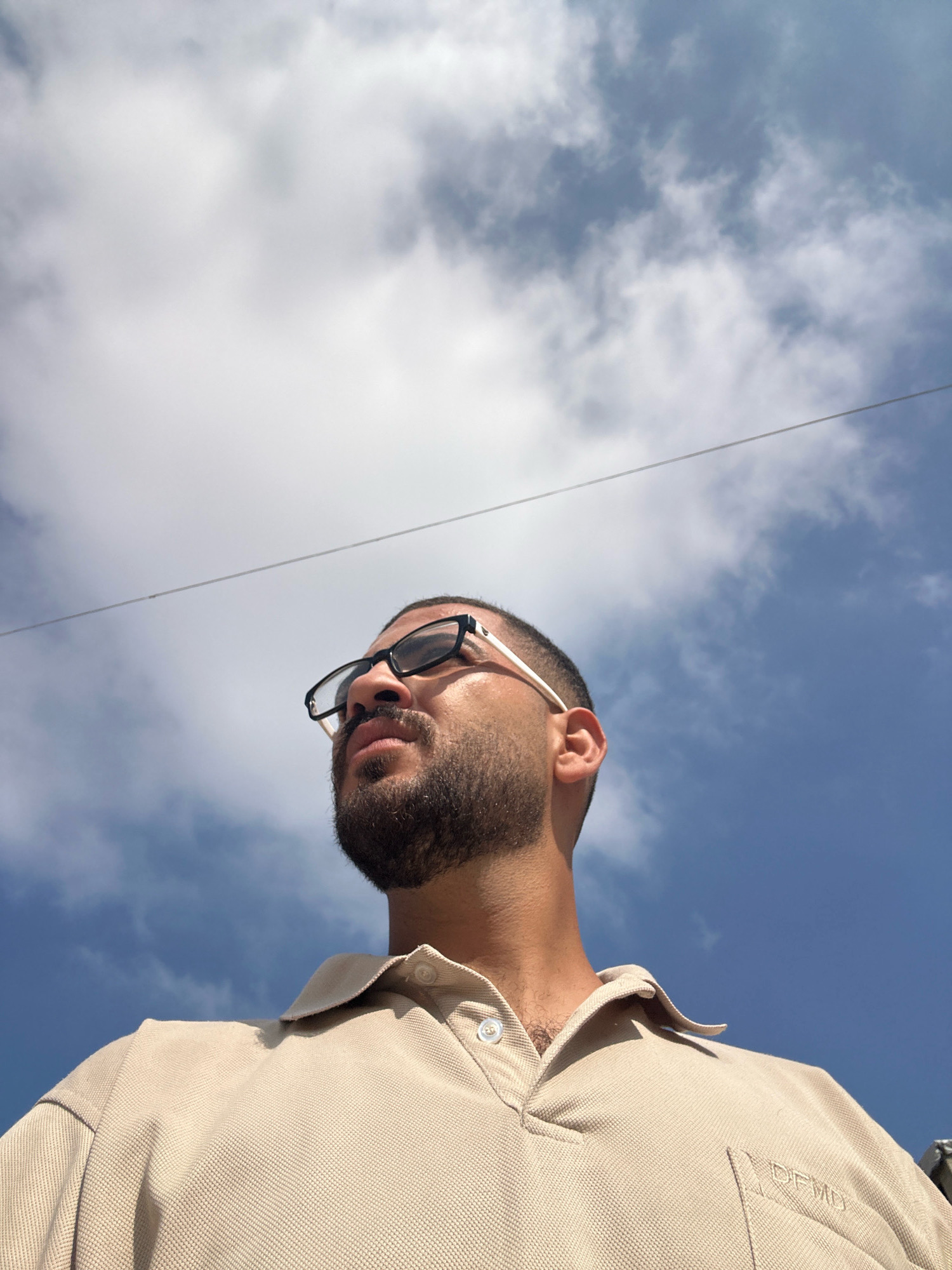 A low-angle outdoor shot shows a man wearing a beige polo shirt and black-rimmed glasses. He has a beard and is looking up and to the left. The background consists of a bright blue sky with white clouds and a thin, dark horizontal line, possibly a wire. The image appears to have been taken in Gaza City, Palestinian Territories, on a sunny day.