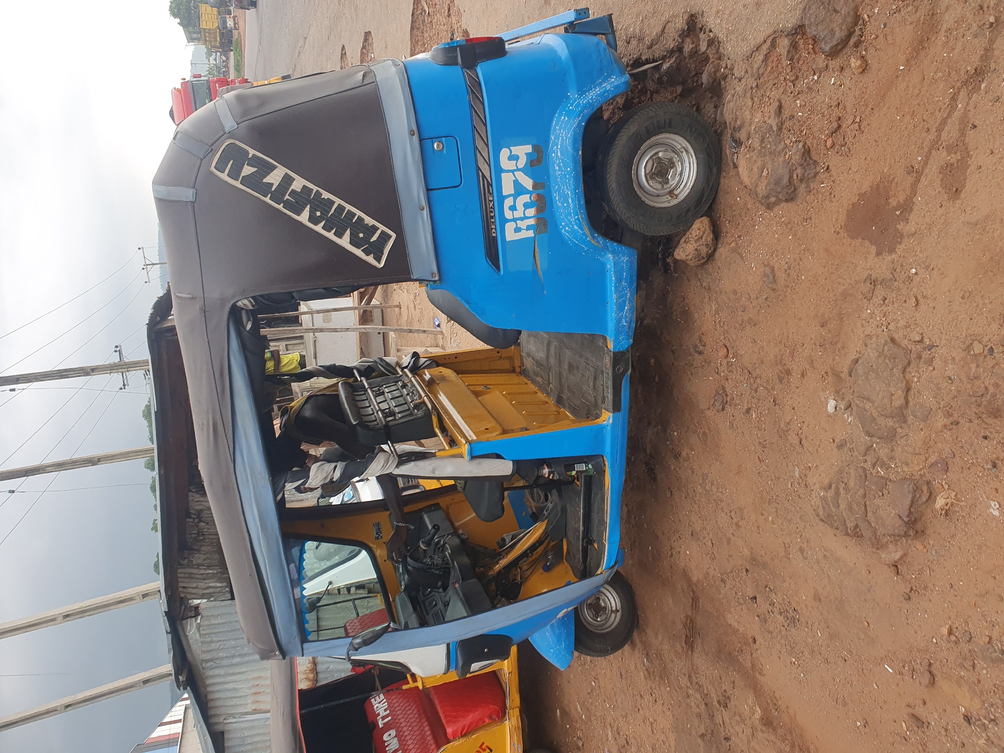 In Lokoja, Nigeria, a blue and yellow auto-rickshaw, identified by the license plate "R679" and the word "DELUXE" on its side, is parked on a dirt road with visible potholes and scattered rocks. The auto-rickshaw features a black canvas roof with a diagonal white graphic that reads "YAMAHIZU." A person, partially visible and wearing a yellow garment, is seated inside the auto-rickshaw, near the steering wheel. Another auto-rickshaw, predominantly yellow with red seating, is parked to the left of the blue one. In the background, a corrugated metal roof structure is visible, and beyond that, a red truck and other vehicles suggest a roadside or market area. The sky is overcast, indicating a cloudy day.