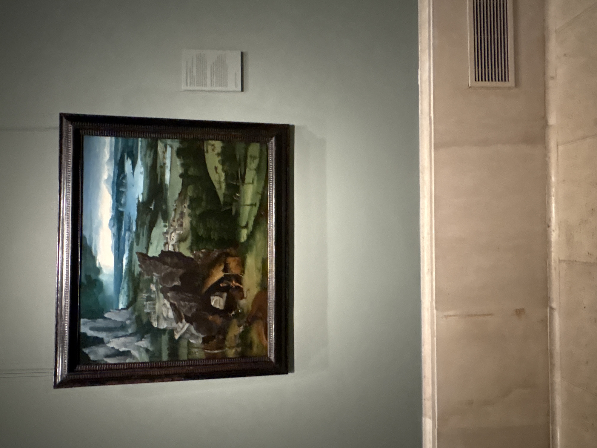 The image shows a painting hanging on a light green wall in what appears to be a gallery or museum setting. Above the painting is a white rectangular sign or placard with text. To the right of the painting is a section of wall, likely part of a doorframe or architectural feature, with a vertical ventilation grille near the top. The wall is a lighter shade, possibly beige or off-white. The painting itself is a landscape scene with blue and grey cliffs, mountains, and water in the background, transitioning to green trees, plants, and brown figures in the foreground. The painting has a dark ornate frame. The gallery's location is Madrid, Spain.