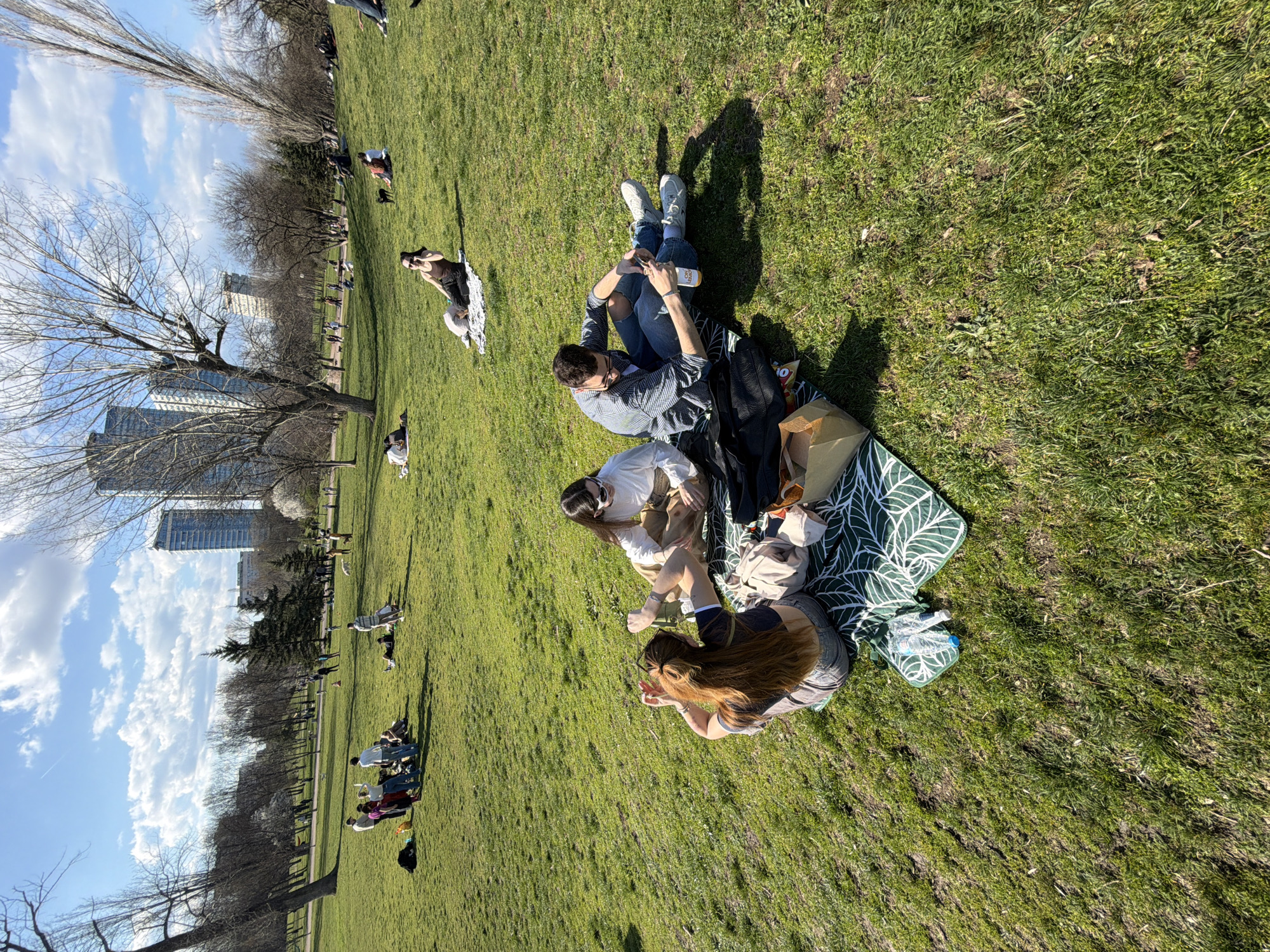 The image shows a grassy area with several groups of people relaxing on a sunny day. In the foreground, four people are sitting on a patterned picnic blanket. One person is lying down, two are sitting and interacting, and one is sitting with legs outstretched, looking at a phone or other handheld device. Around them are a few bags, bottles, and other items. In the background, other people are scattered across the grass, some sitting and some walking. There are trees lining the left side of the image, and in the distance are buildings, suggesting this is a park in an urban area. Based on the architectural style of the distant buildings and the overall atmosphere, the location is likely a park in Sofia, Bulgaria.