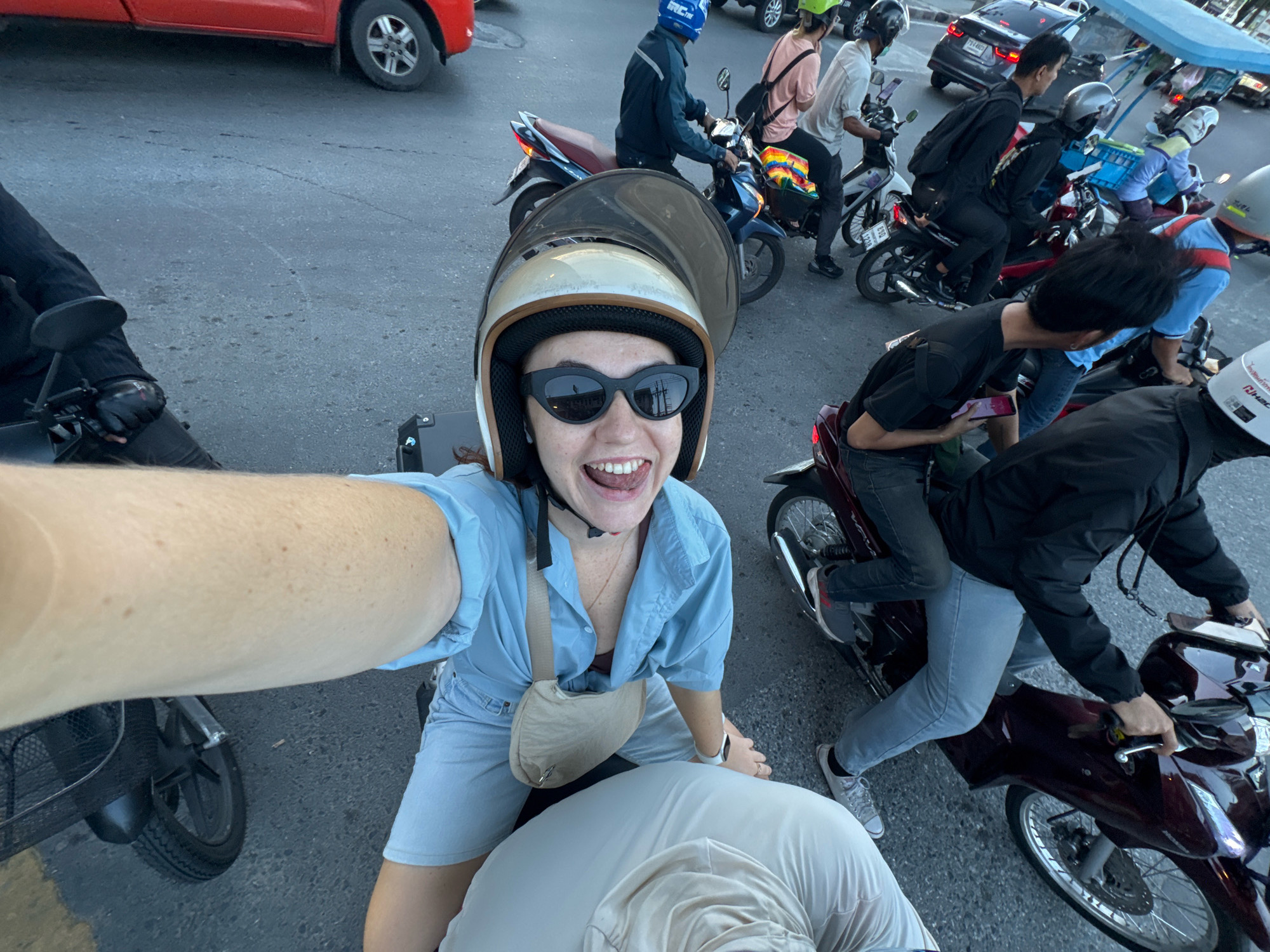 In Samrong Nuea Subdistrict Municipality, Thailand, a woman with a helmet and sunglasses rides on the back of a scooter in traffic. She is wearing a blue button-down shirt, shorts, and a small beige purse across her chest. A man in a black jacket and grey pants drives the scooter. They are in a line of other scooters and a red car on a paved road. Other individuals are also visible on motorcycles in the background.