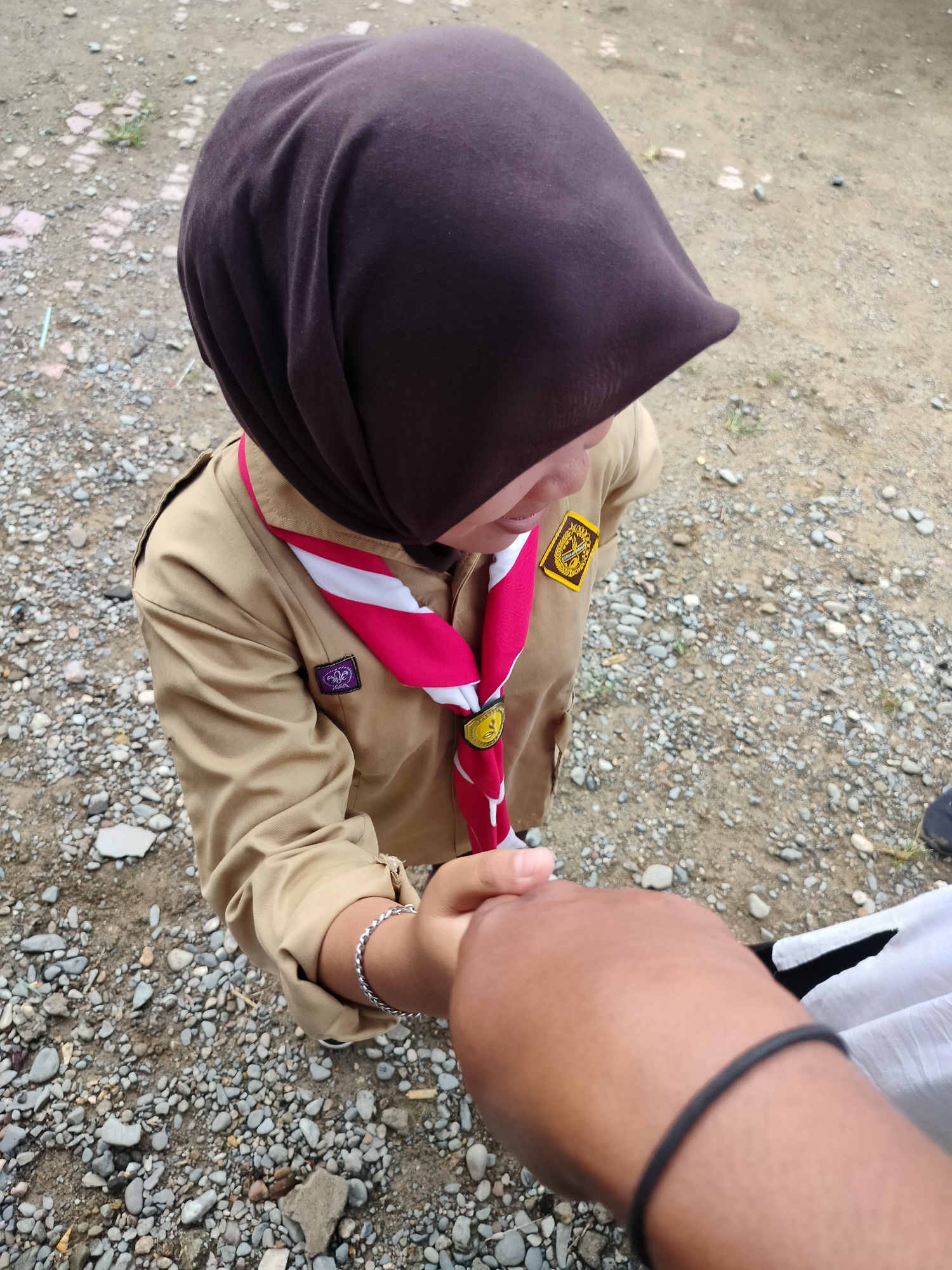 In Lawebekung, Indonesia, a person wearing a brown scout uniform and a dark brown hijab is holding hands with another person. The scout has a red and white scarf around their neck and several badges on their uniform. They are standing on a ground covered with small rocks and pebbles. The overall scene suggests an outdoor setting, possibly a training or gathering for scouts.