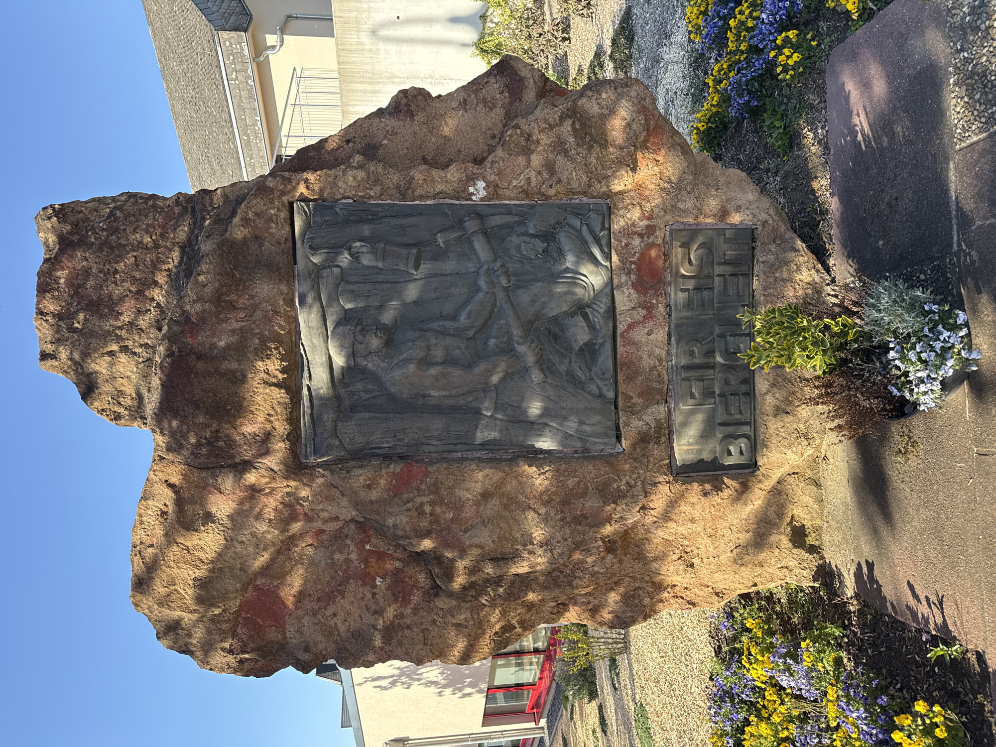 This media file captures an outdoor monument, likely a memorial, under a clear blue sky. The central subject is a large, rugged boulder of reddish-brown rock, featuring two embedded metal plaques.
The larger plaque, situated on the left side of the boulder, is a rectangular bronze bas-relief sculpture. It depicts several human figures, appearing to be miners. One figure in the foreground is shown kneeling or bending, wielding a pickaxe. Other figures stand behind, also engaged in what seems to be manual labor, possibly within a mine or quarry setting.
To the right of the bas-relief, a smaller, rectangular metal plaque is embedded vertically into the rock. This plaque features raised capital letters. The clearly visible text reads "BEFREIT", which is German for "Liberated".
The monument is set amidst landscaping that includes vibrant beds of yellow and purple pansy-like flowers, particularly prominent on the right side of the image. There are also patches of smaller green plants, gravel, and paved areas around the base of the boulder.
In the background, under the bright blue sky, parts of buildings are visible. One building features a dark-tiled roof and light-colored walls, while another has a more modern appearance with noticeable red window frames.
The scene is illuminated by strong, direct sunlight, indicating a clear day. This lighting creates distinct shadows on the monument and the surrounding ground. Given the location context of Pétange, Luxembourg, and the visible text "BEFREIT", the monument likely commemorates a significant liberation event or aspects of the region's industrial history, possibly related to mining.