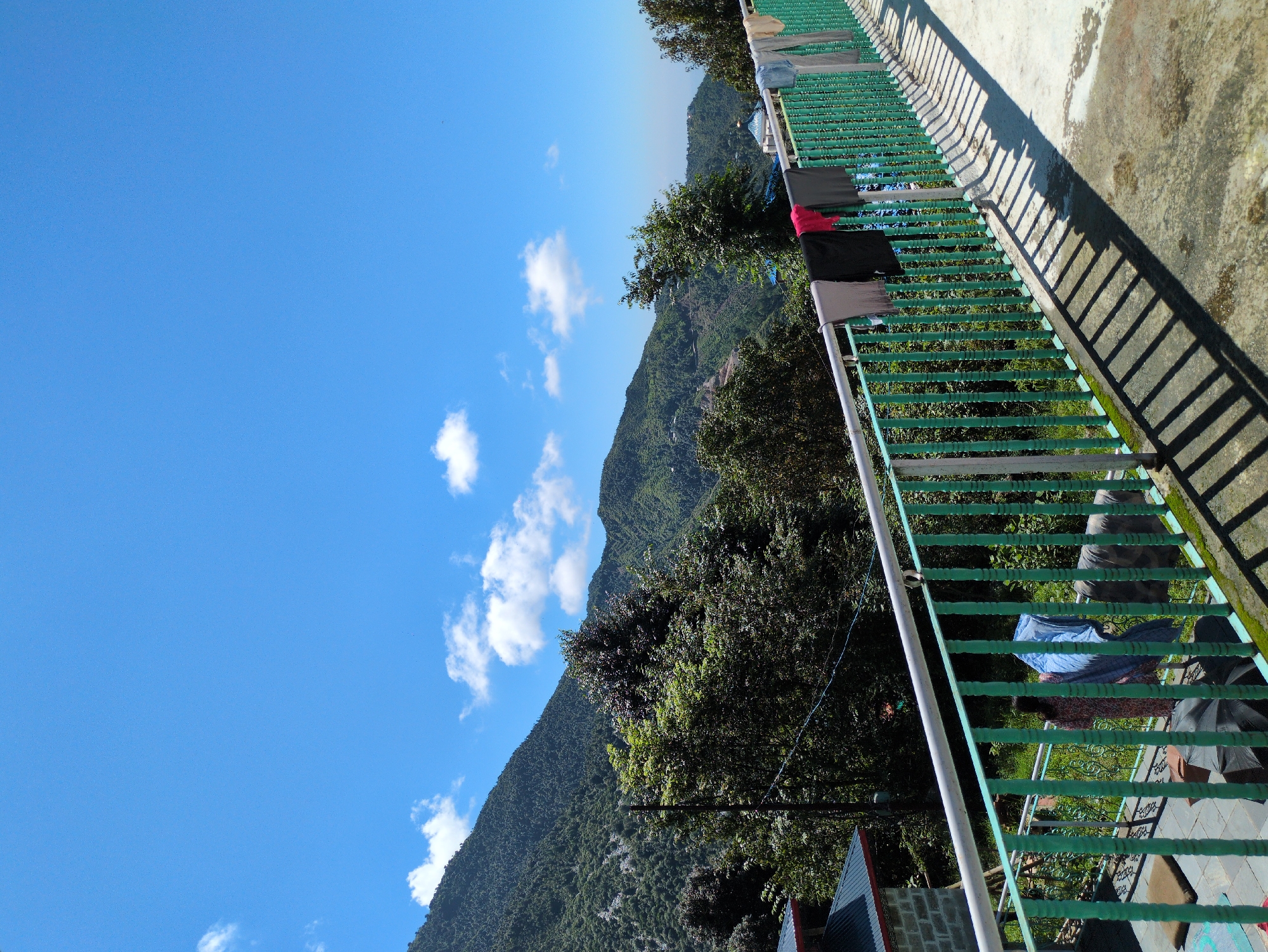 The image depicts a scenic view of a mountainous region with a clear blue sky and scattered white clouds. In the foreground, a railing with green painted vertical slats and a white horizontal bar runs across the frame. Several pieces of laundry are hanging from the railing, including shirts and pants in various colors. The ground in the foreground is a concrete surface with distinct shadows cast by the railing. A building with a grey brick facade and a dark tiled roof is partially visible on the left. The background is dominated by lush green hills and mountains, covered in dense vegetation and trees. Some structures, possibly houses, are scattered along the hillsides. The overall impression is a peaceful, outdoor scene in a hilly area, likely in a rural or semi-rural setting in Haini, India.