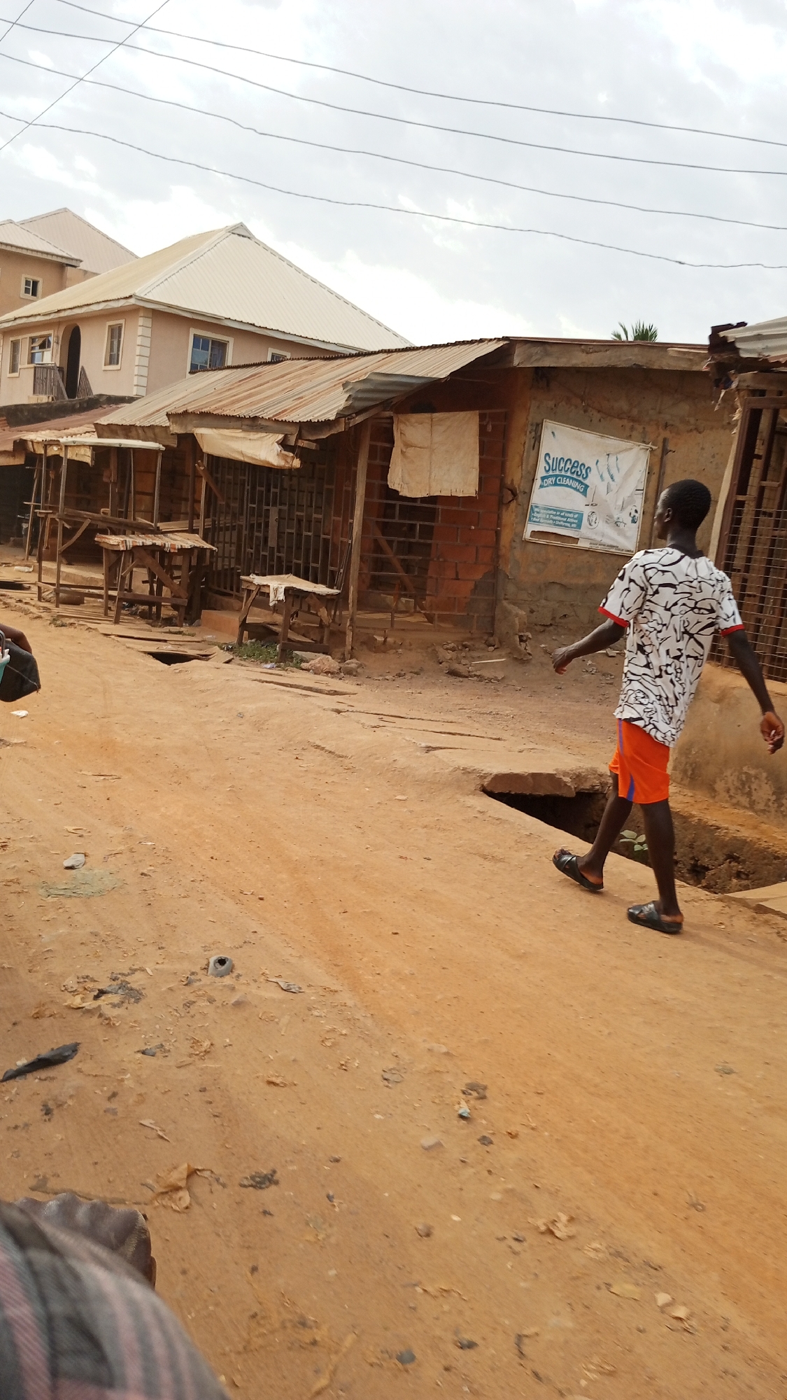 The image captures a street scene in Ajaokuta, Nigeria, under an overcast sky during daytime. The primary subject is a young man walking across an unpaved, reddish-brown dirt road, moving from left to right away from the camera. He wears a white t-shirt with a black abstract pattern and red trim on the sleeves, orange shorts, and black sandals. Part of another person, possibly the photographer, is visible in the bottom left corner, wearing a striped garment.
The setting features a mix of buildings. In the background, there's a more modern, two-story house with a light-colored exterior and a light-colored tiled roof. In the midground, a row of smaller, single-story structures lines the street. These appear to be market stalls or shops, characterized by their rustic construction with brick walls, wooden frames, and rusty corrugated metal roofs. Several empty wooden tables or stands are positioned in front of these shops. Power lines crisscross the sky above the buildings.
Visible infrastructure includes several open drain covers or holes along the edge of the dirt road. A white banner attached to one of the shops prominently displays "Success DRY CLEANING" in blue text, with smaller, illegible text below it. The road itself is uneven and scattered with small debris. The overall atmosphere suggests a quiet moment in a local commercial or residential area.