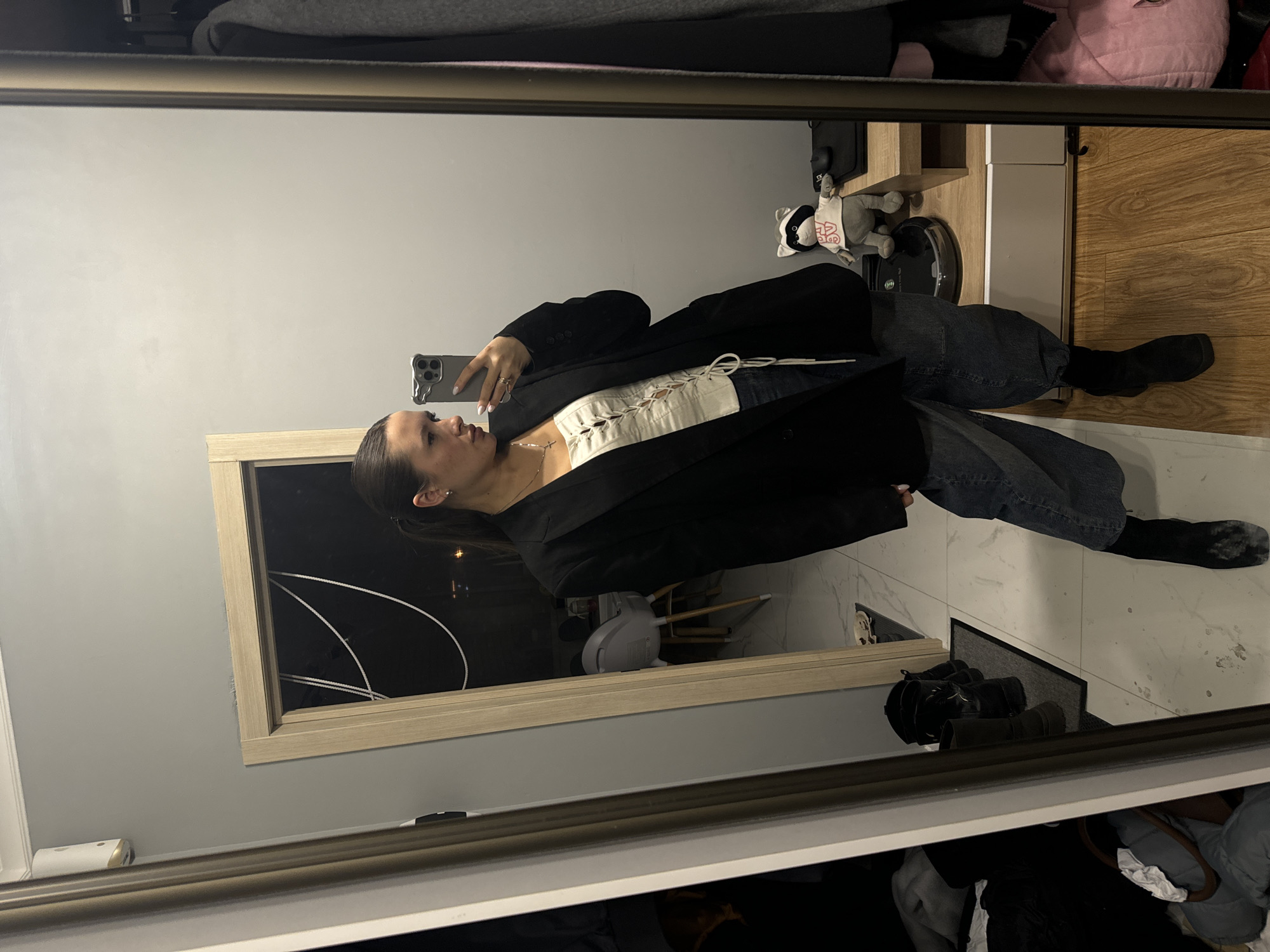 A woman is visible in a mirror taking a selfie with her phone. She is wearing a white corset-style top, a black blazer, jeans, and black boots. The background includes part of a room, what appears to be a hallway with a white marble tiled floor, and a window reflecting an outside night scene. A vacuum cleaner and a plush toy are visible on a wooden surface in the background. The overall environment appears to be an indoor residential space. There's no clear evidence indicating this is in the city of Odintsovo, Russia.