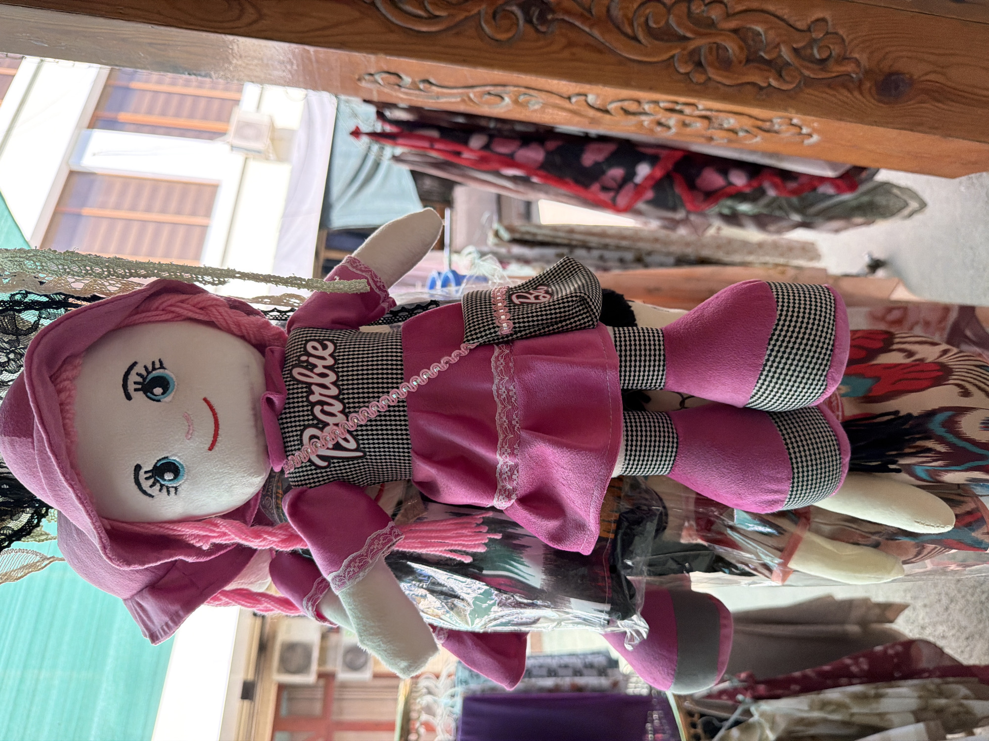 An eye-level, medium shot captures a plush doll, likely a Barbie doll, hanging by a string in an outdoor marketplace. The doll is primarily pink, with a pink hat, pink dress adorned with a black and white checkered pattern and the word "Barbie" in cursive, and pink boots. It has blue eyes, black eyelashes, and rosy cheeks. A small black and white checkered purse with a pink strap hangs from its shoulder. The doll is wrapped in a clear plastic bag, possibly for protection.

In the background, a variety of fabrics and garments are displayed, including scarves and dresses with colorful patterns. A wooden post with intricate carvings stands to the right of the doll. The overall environment suggests a market stall in a city. The presence of air conditioning units on a building in the background indicates a semi-urban or urban setting. The lighting is bright, consistent with daylight. The camera focuses on the doll, with the background elements slightly blurred. The scene is set in the city of Zangiata, Uzbekistan.