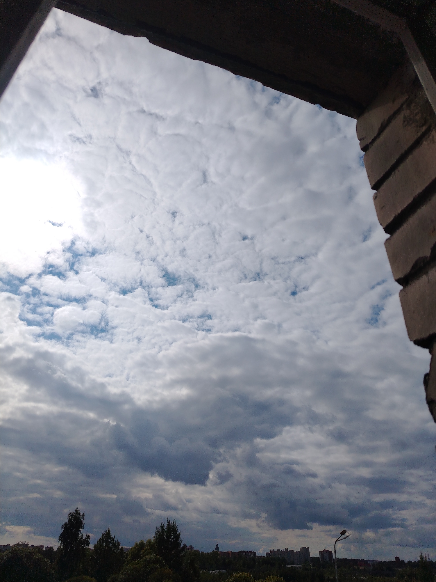 This image depicts a view of the sky through an opening, possibly a window or doorway, framed by dark, rough material at the top and a brick structure on the right. The sky is filled with a variety of clouds, ranging from bright white, puffy cumulus clouds on the left, illuminated by the sun, to darker, more ominous clouds towards the bottom and right. The sun is visible as a bright, hazy area on the upper left.
Below the sky, a silhouetted treeline forms the horizon, with a few taller trees standing out. In the distance, the indistinct shapes of buildings and a streetlamp suggest an urban or suburban setting. The overall impression is that of an overcast or partly cloudy day, viewed from a sheltered vantage point overlooking a cityscape.
While the image provides a general sense of a city, it does not contain specific landmarks that would allow for precise identification of Vyborg, Russia. The limited details visible in the distance are too generic to pinpoint a specific location within the city.
