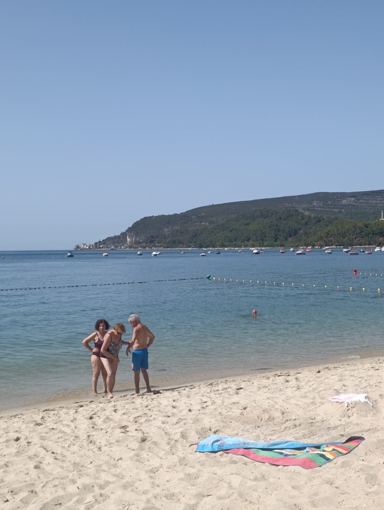 The image depicts a sunny day at a beach in Setúbal, Portugal, likely facing the Sado River estuary. In the foreground, a sandy beach stretches out, with two towels laid out on the sand. One towel is a colorful striped towel, and the other is a smaller white towel with a pink edge. Closer to the water, three people stand near the shoreline. Two women, dressed in swimsuits, and one man in blue swim trunks are engaged in a conversation. The water is calm and blue, with a few boats anchored in the distance. Beyond the water, a tree-covered hillside rises, with some buildings visible at its base, characteristic of the Arrábida Natural Park region near Setúbal. The sky is clear and a pale blue.