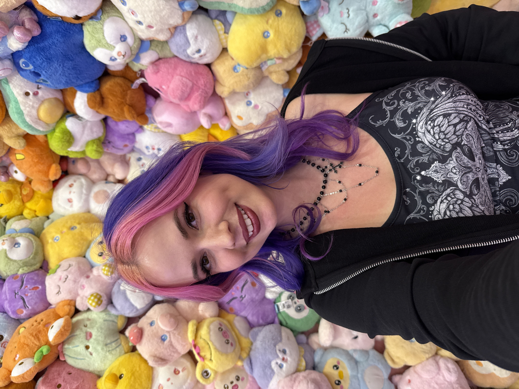 A woman with pink and purple hair is lying among many colorful, stuffed animal toys. She is wearing a black zip-up jacket over a patterned black and gray top and a silver cross necklace. The setting resembles an indoor location, possibly a room filled with plush toys, and the absence of recognizable landmarks makes determining if the location is Madrid, Spain, impossible without additional information.